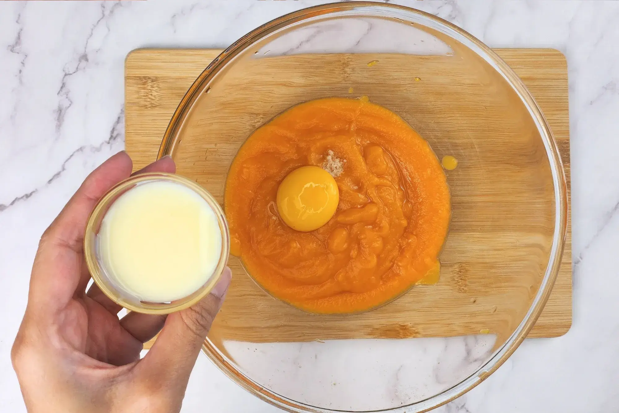 A hand is holding a bowl of condensed milk above a mixing bowl.