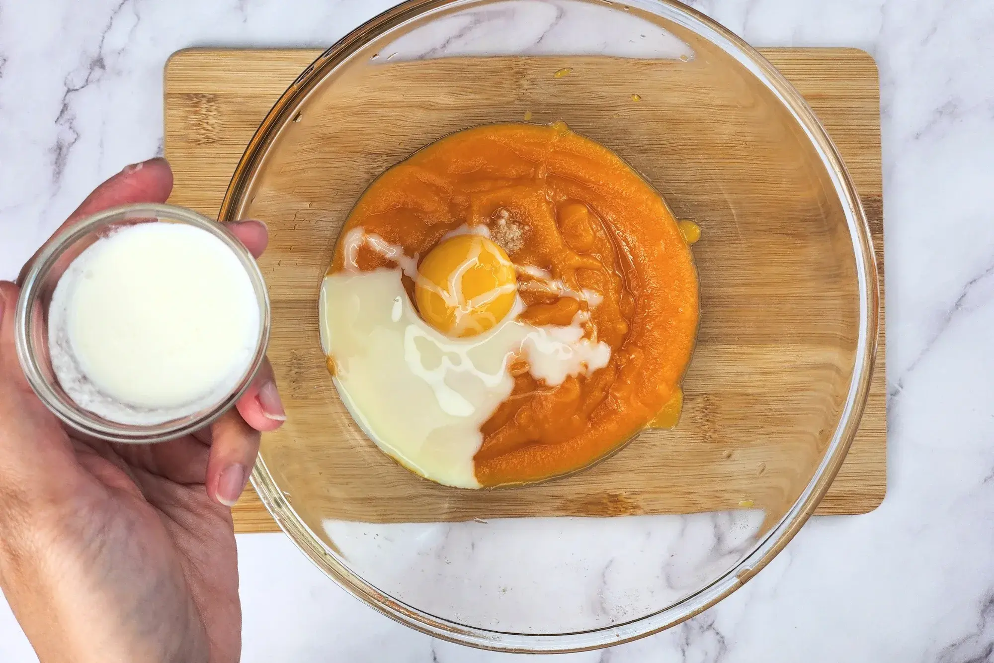 A hand is holding a bowl of heavy cream above a mixing bowl.