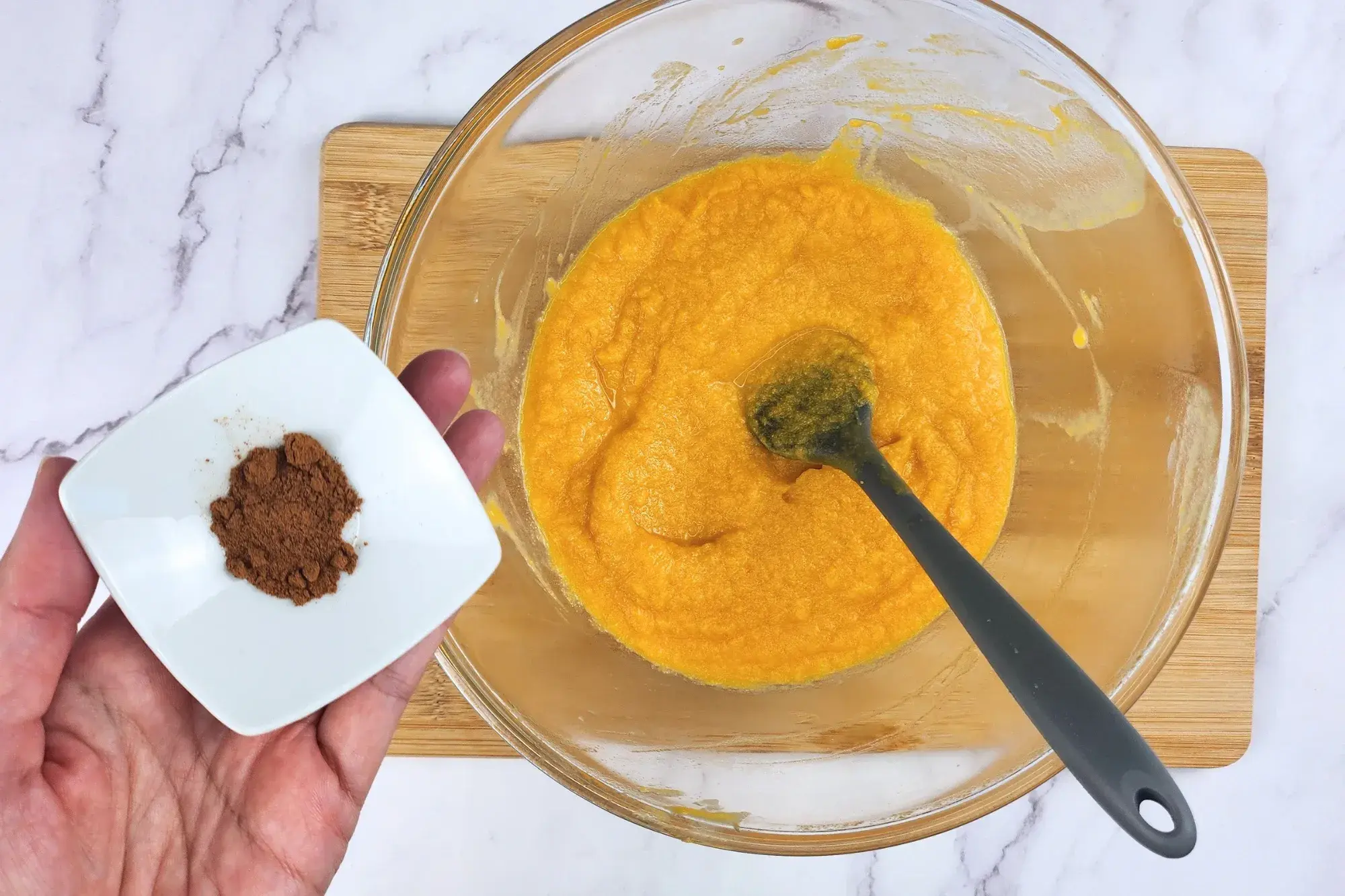 A hand is holding a bowl of cinnamon powder above a mixing bowl.