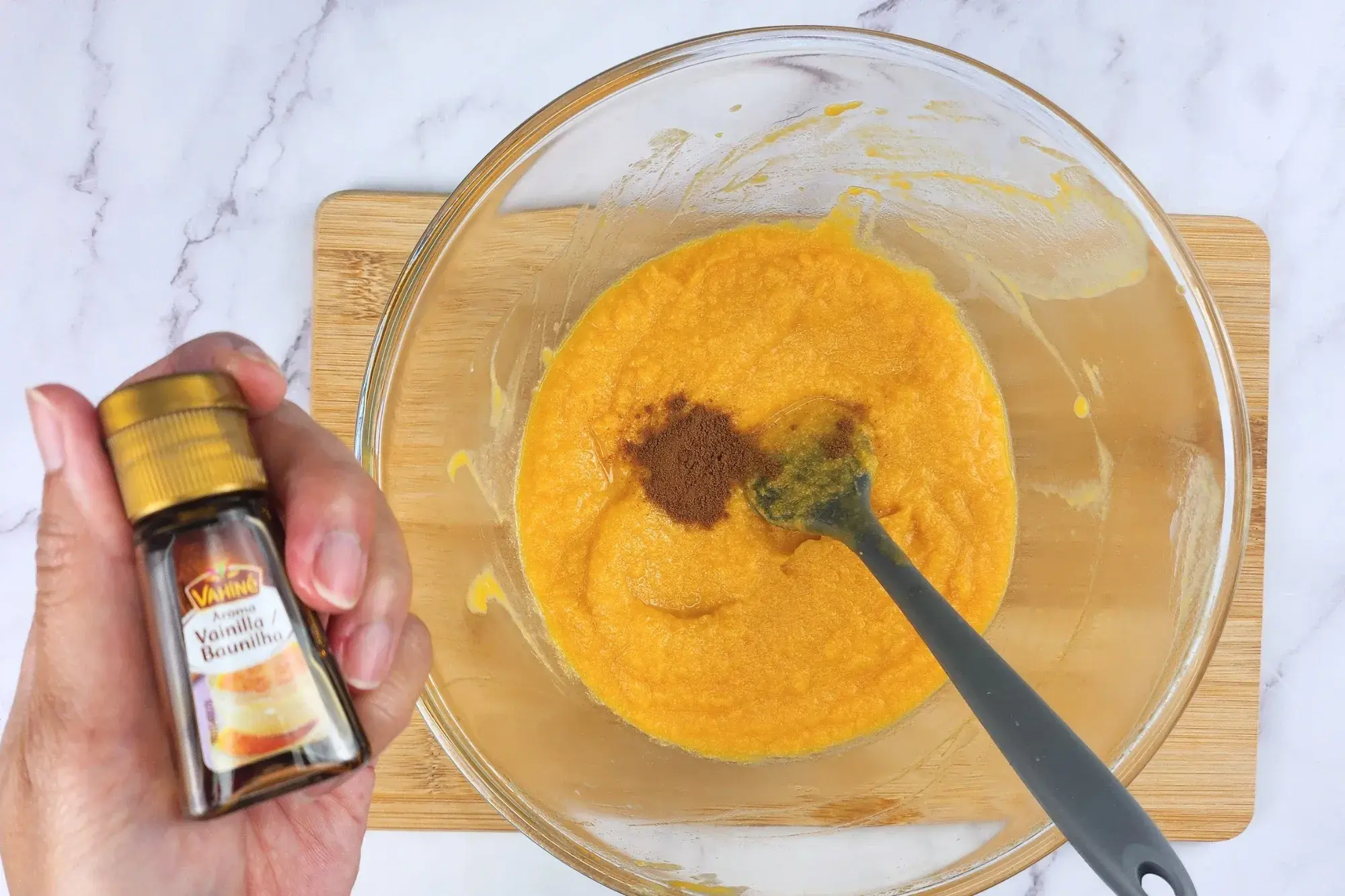 A hand is holding a bottle of vanilla extract above a mixing bowl.