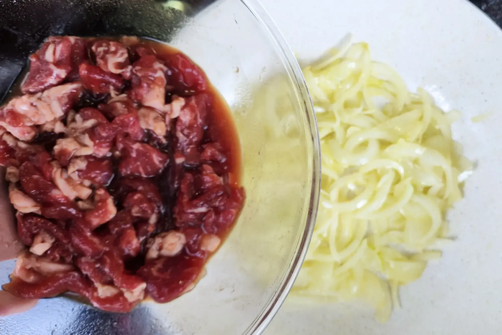 A bowl of seasoned beef sliced above a wok with cooked onion.