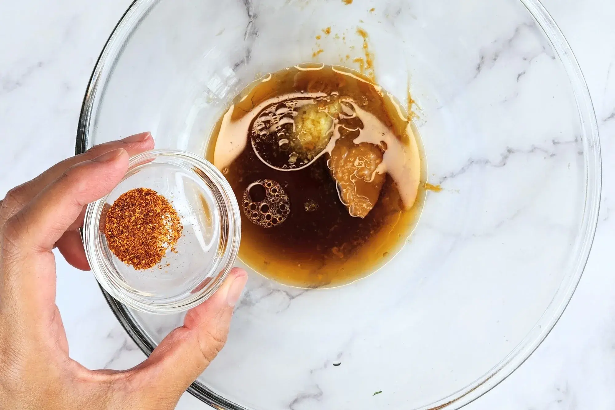 A hand holding a bowl of chili flkes above the dressing bowl