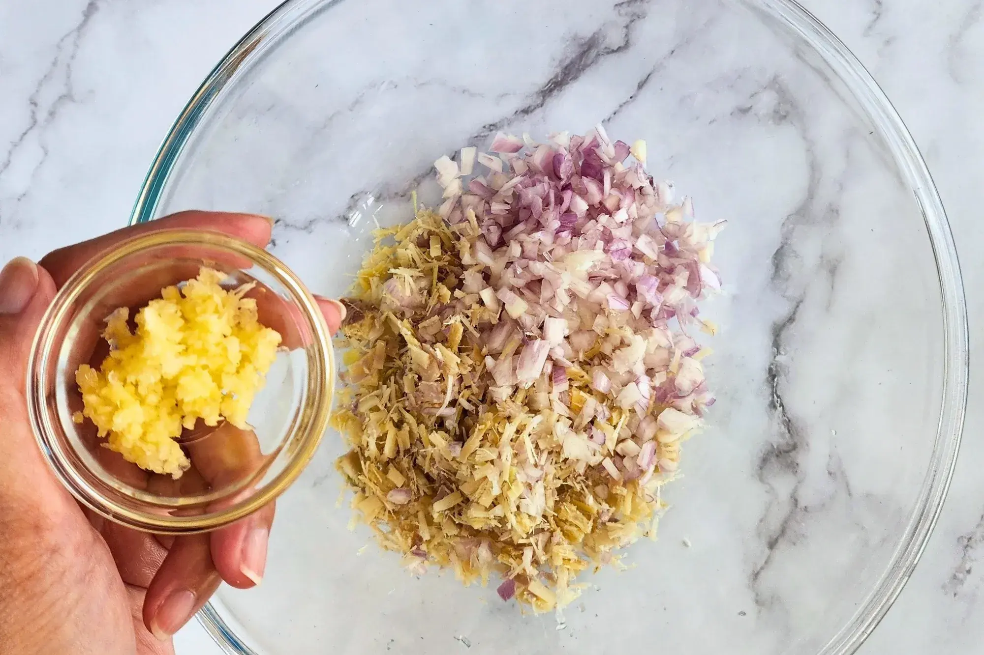 A hand holding a bowl of minced garlic above a mixing bowl