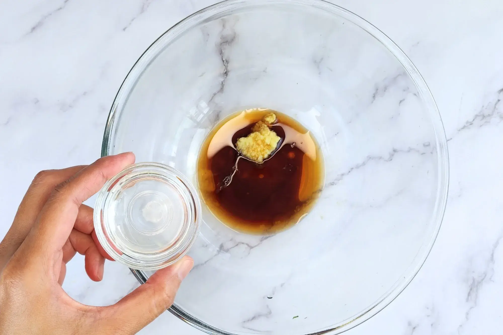A hand holding a bowl of rice vinegar above the dressing bowl
