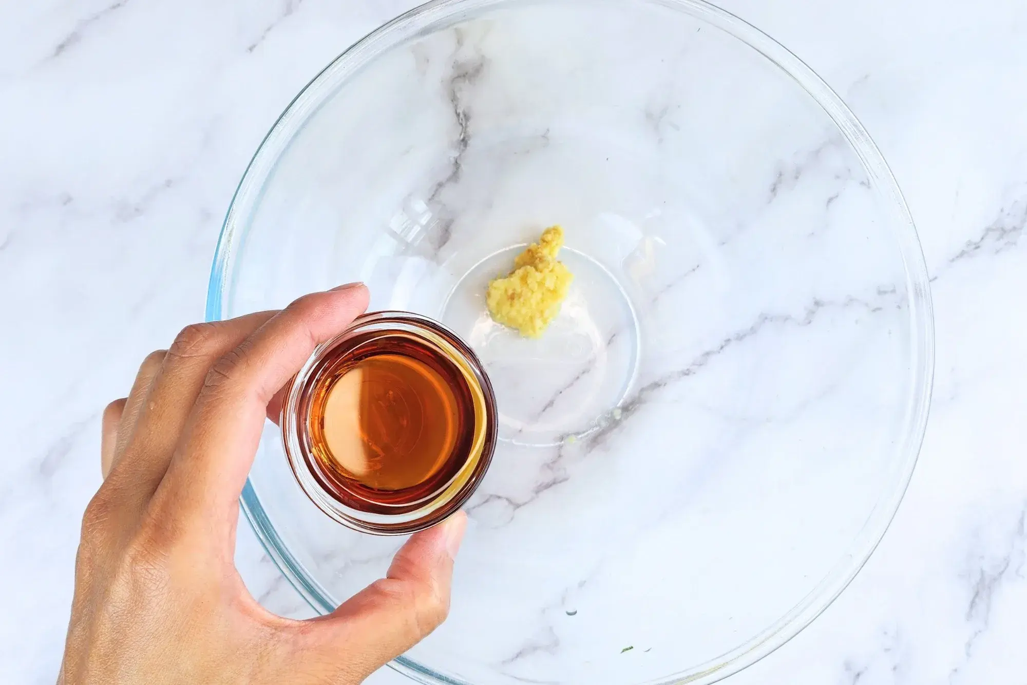 A hand holding a bowl of sesame oil above the dressing bowl
