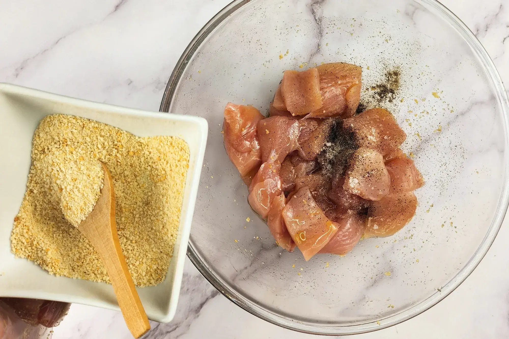 A hand is hold a bowl of breadcrumbs above a mixing bowl with chicken..