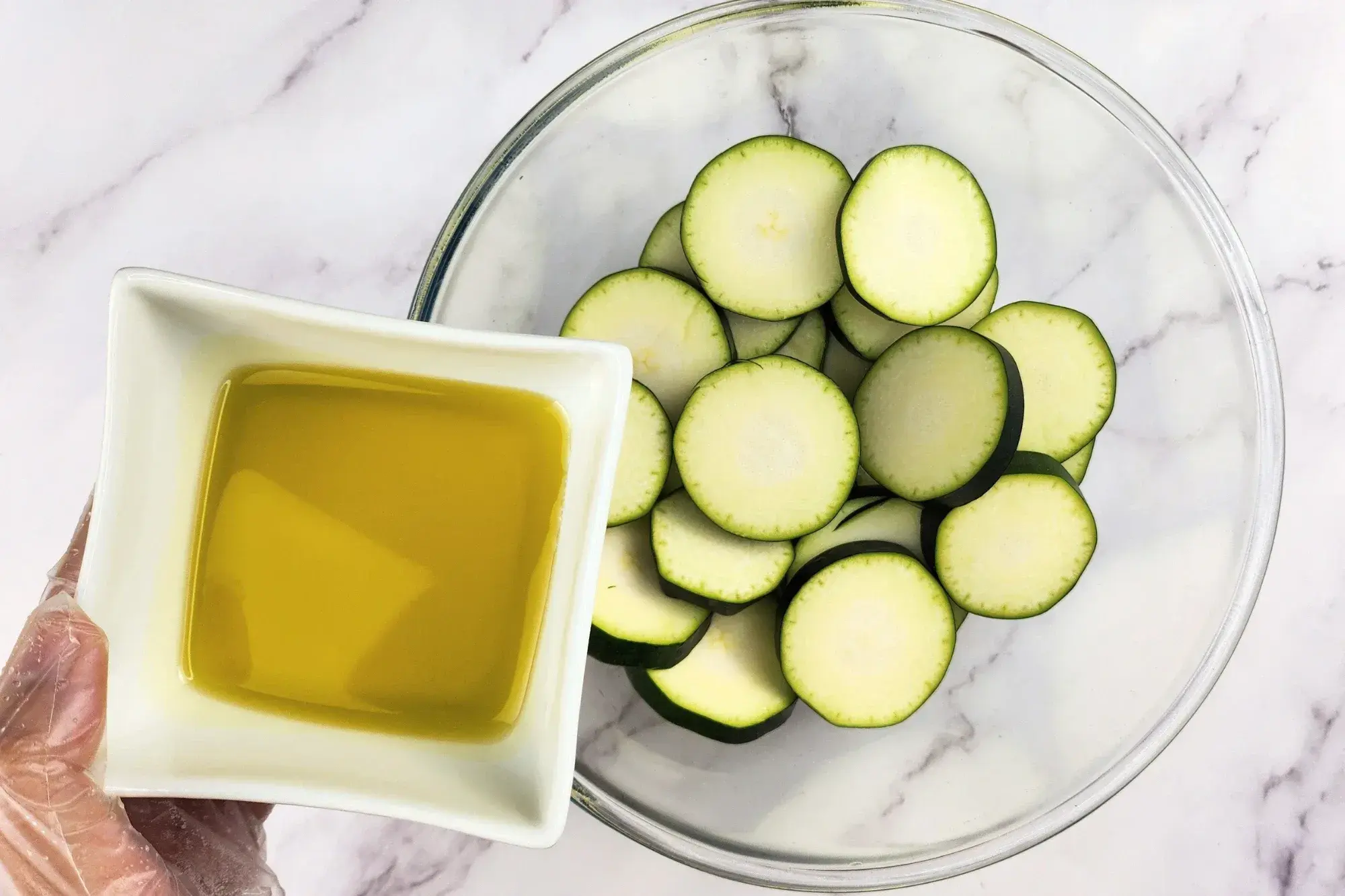 A hand is hold a bowl of olive oil above a mixing bowl with zucchinis.