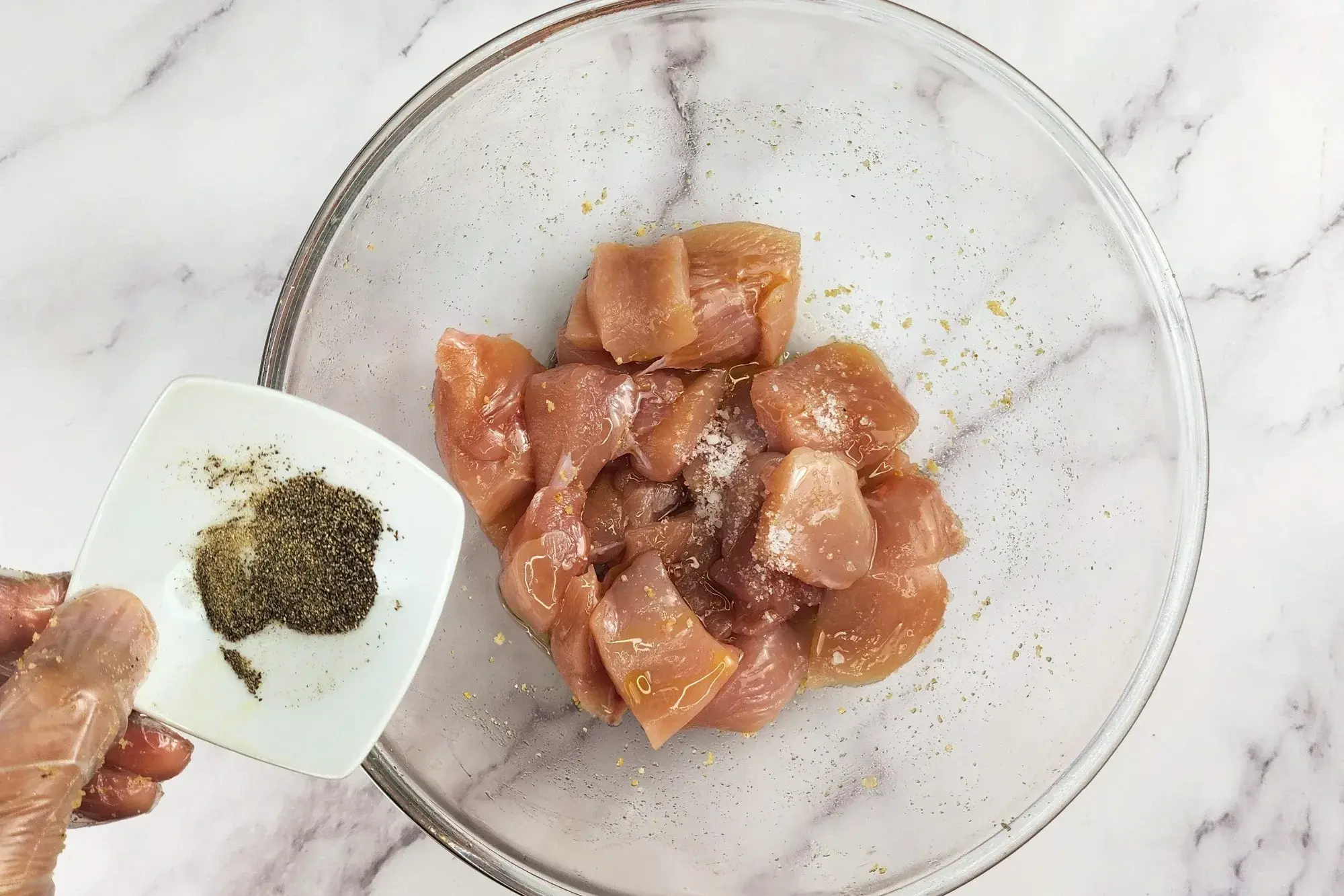 A hand is hold a bowl of pepper above a mixing bowl with chicken.