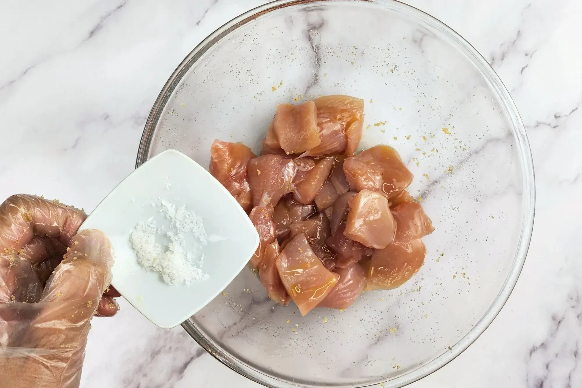A hand is hold a bowl of salt above a mixing bowl with chicken..