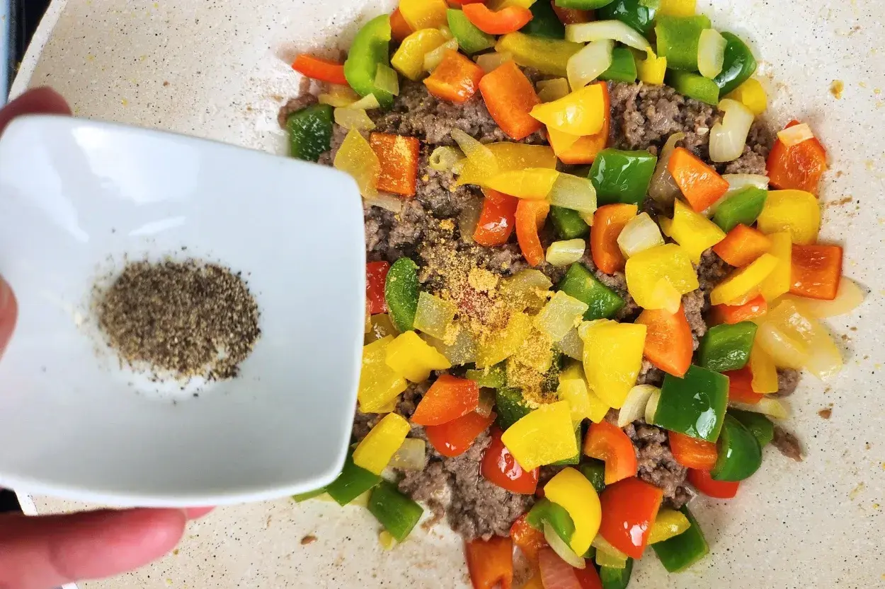 A hand is holding a bowl of black pepper powder above cooked food in a wok.