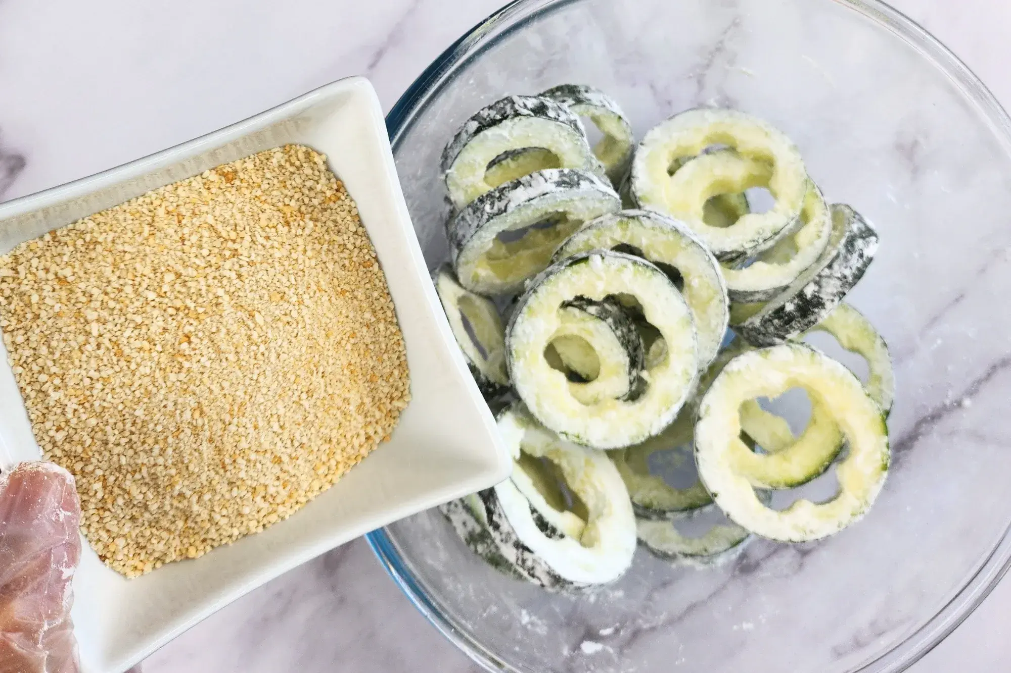 A hand is holding a bowl of breadcrumbs above a bowl of zucchini rings