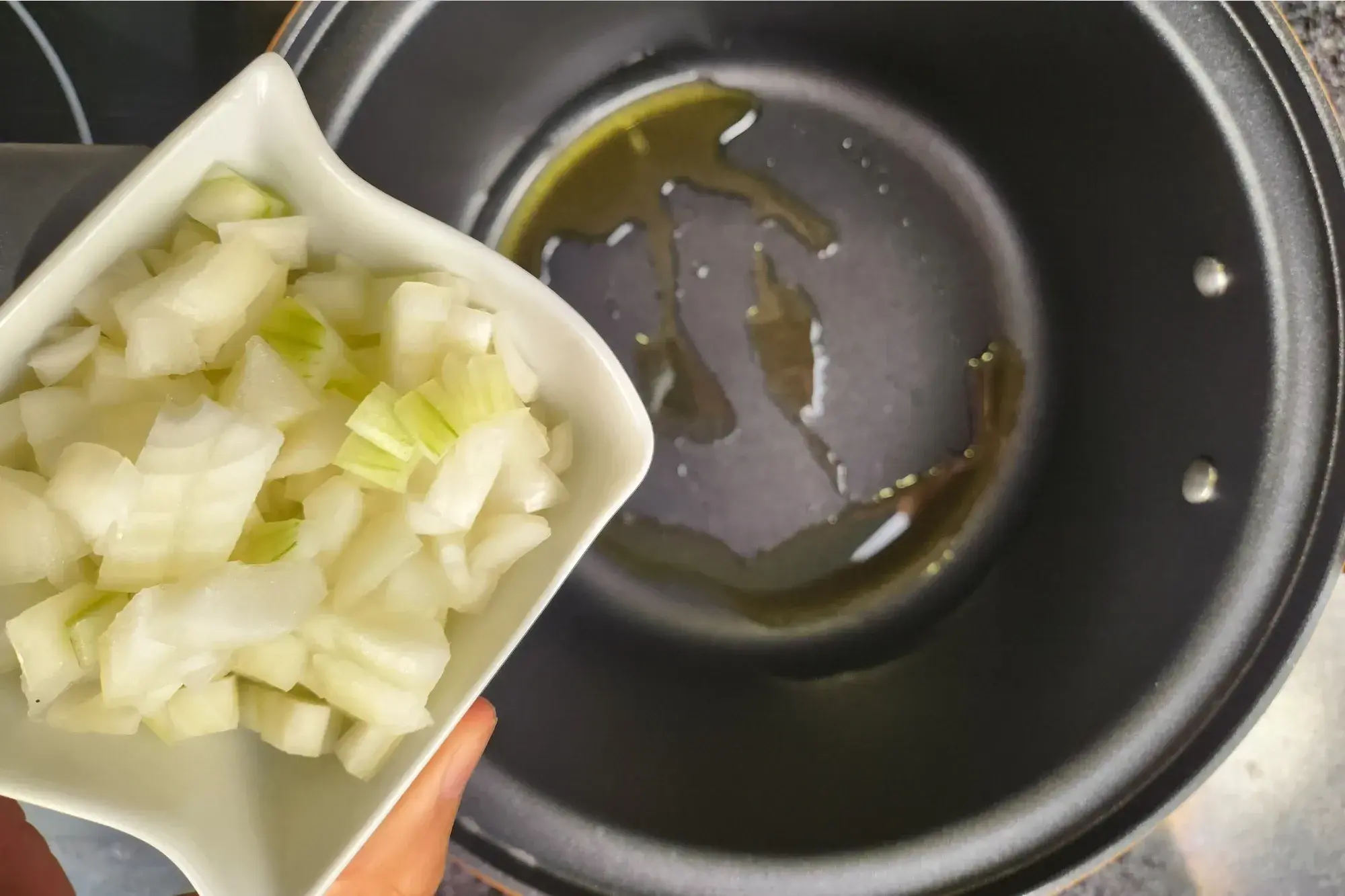 A hand is holding a bowl of chopped onion above a black pot