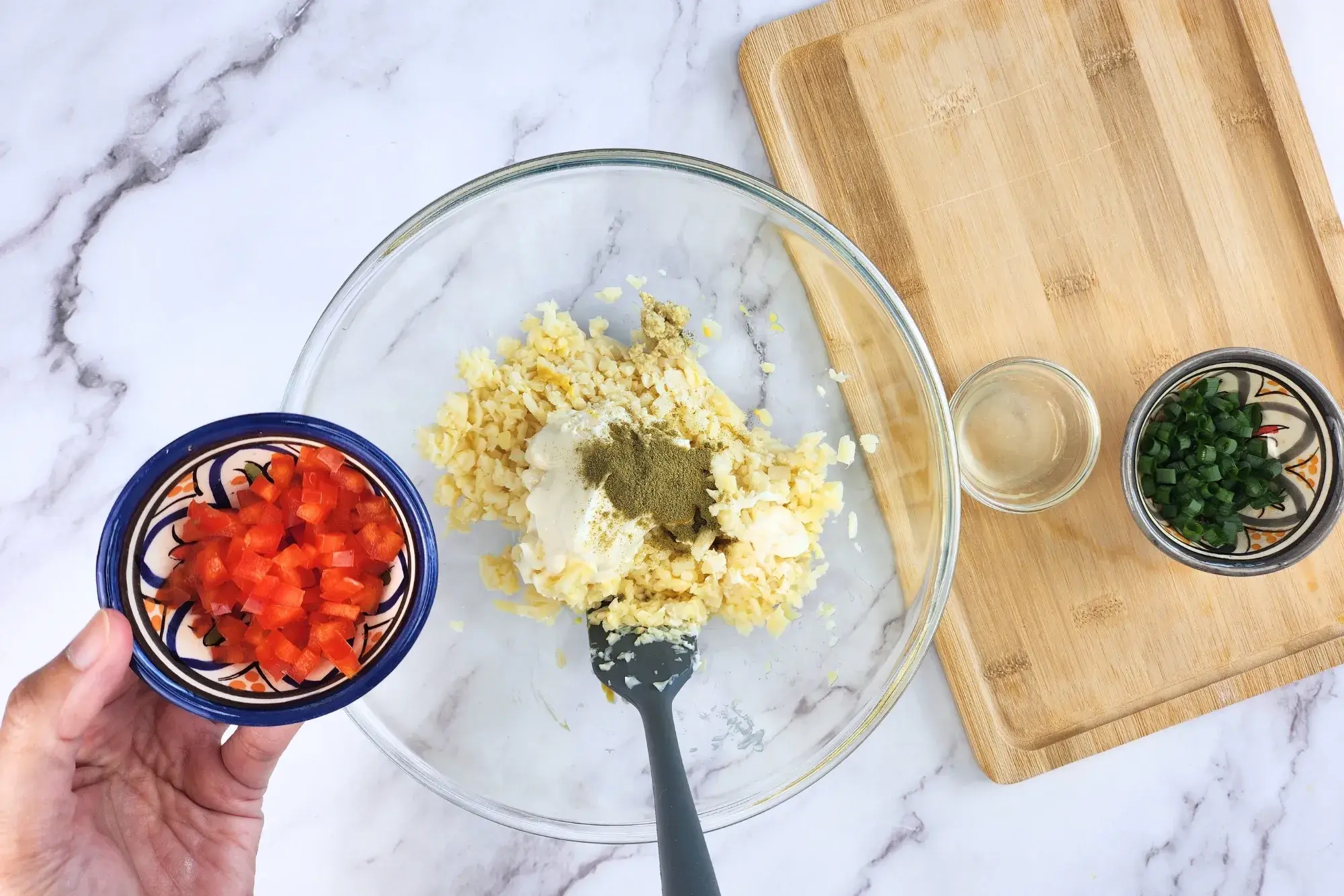 A hand is holding a bowl of chopped red bell pepper above the mixing bowl.