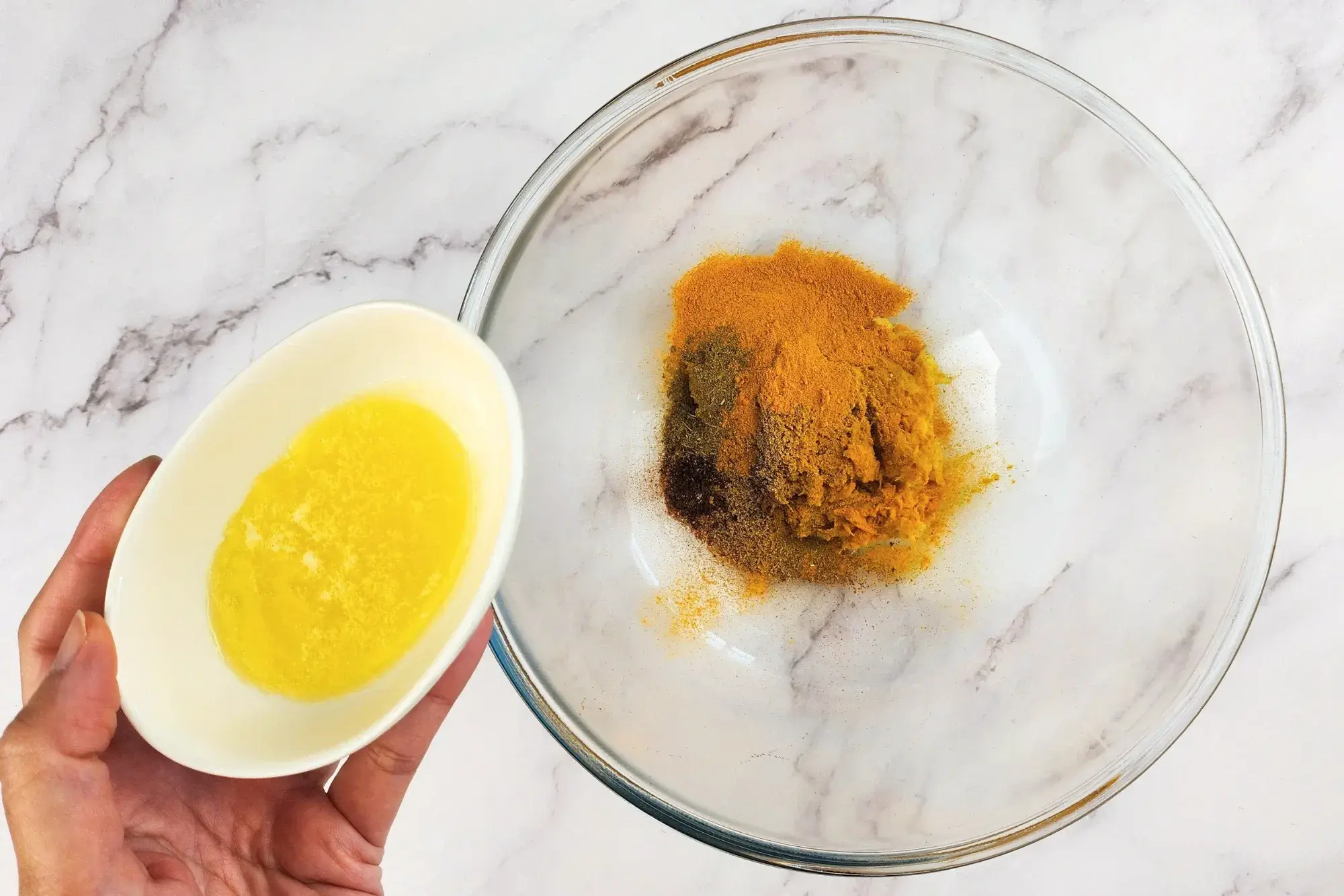 A hand is holding a bowl of melted butter above a mixing bowl with spices.