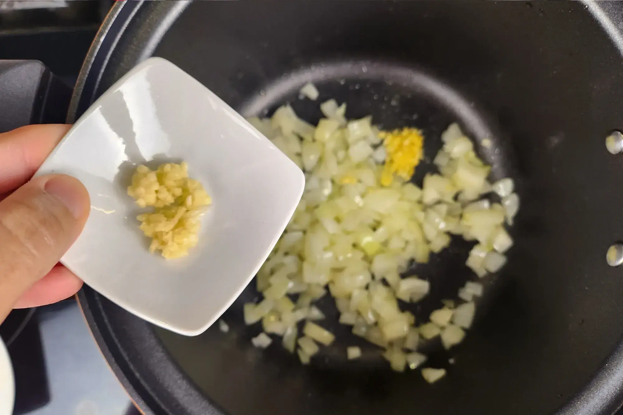 A hand is holding a bowl of minced garlic above a black pot with cooked onion