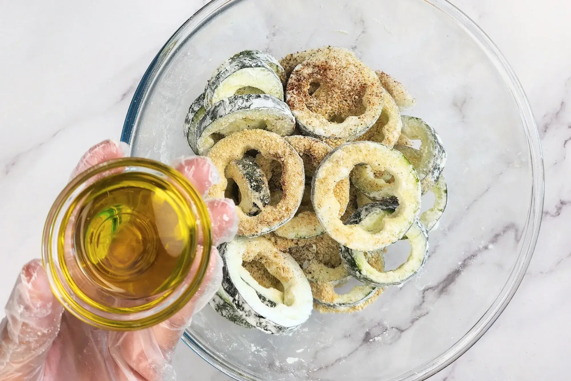 A hand is holding a bowl of oil above a bowl of zucchini rings.