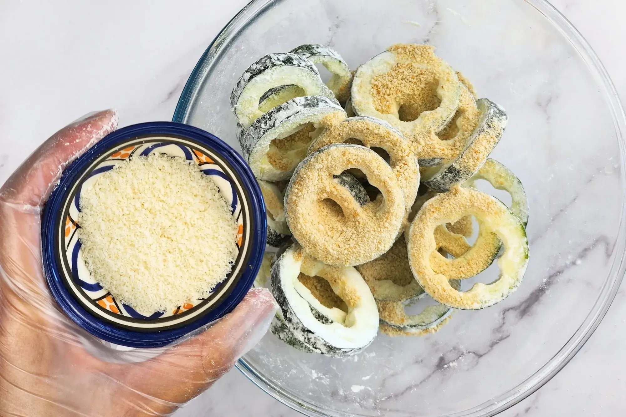 A hand is holding a bowl of parmesan above a bowl of zucchini rings.