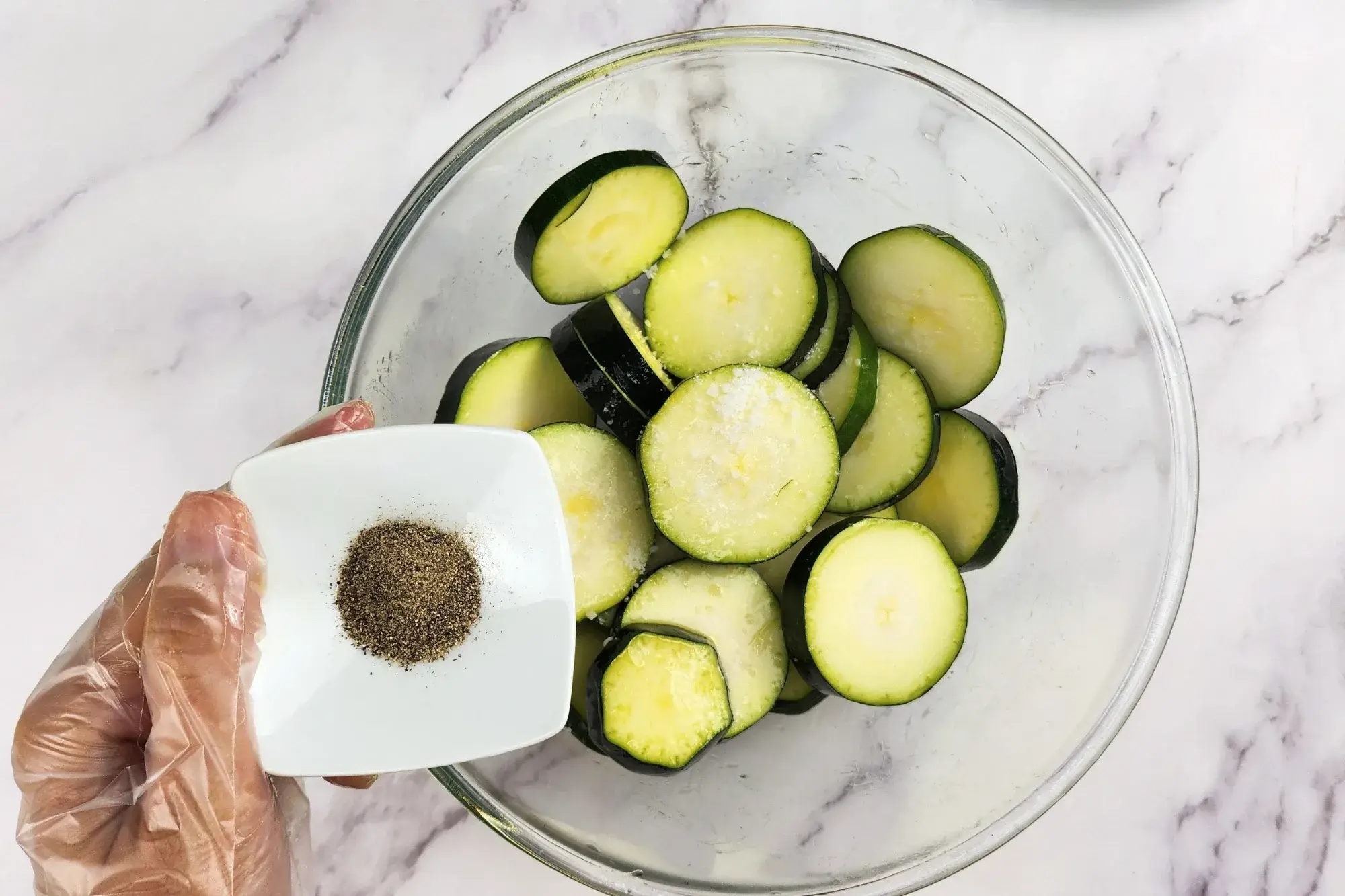 A hand is holding a bowl of pepper above a mixing bowl with zucchinis. 