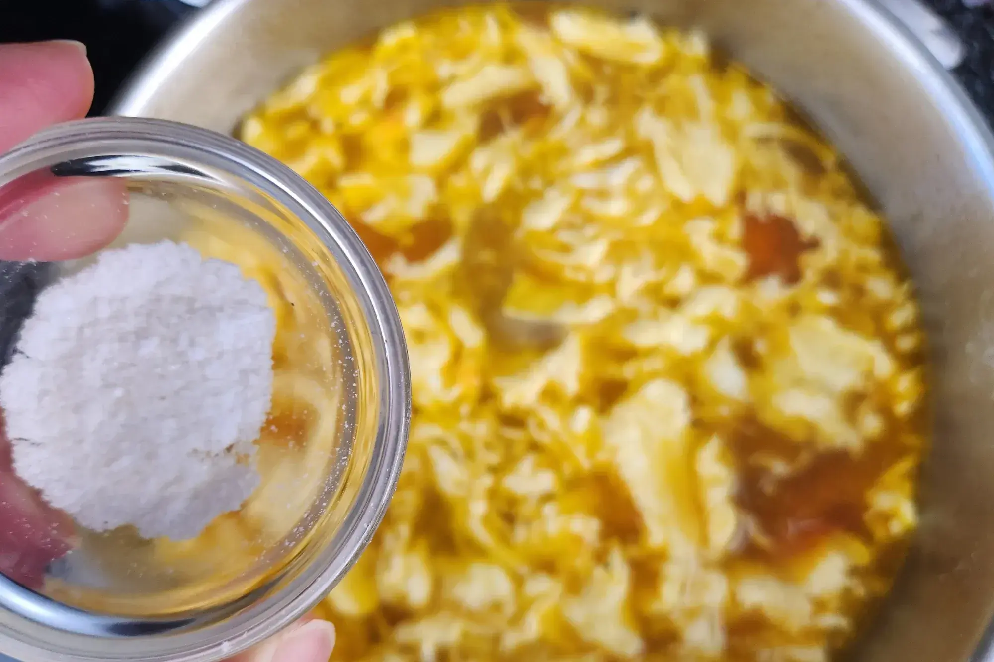 A hand is holding a bowl of salt above of Tomato Egg Drop Soup.