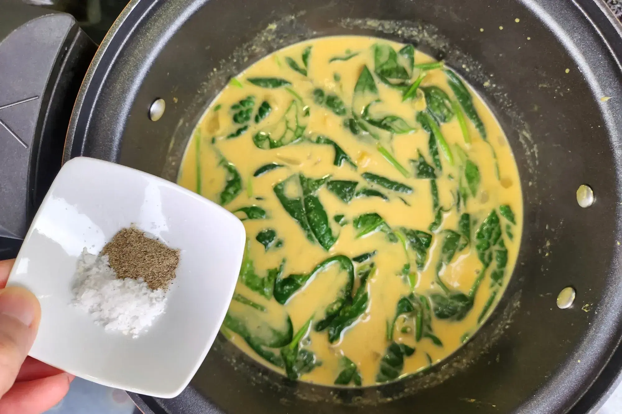 A hand is holding a bowl of salt and black pepper above a pot