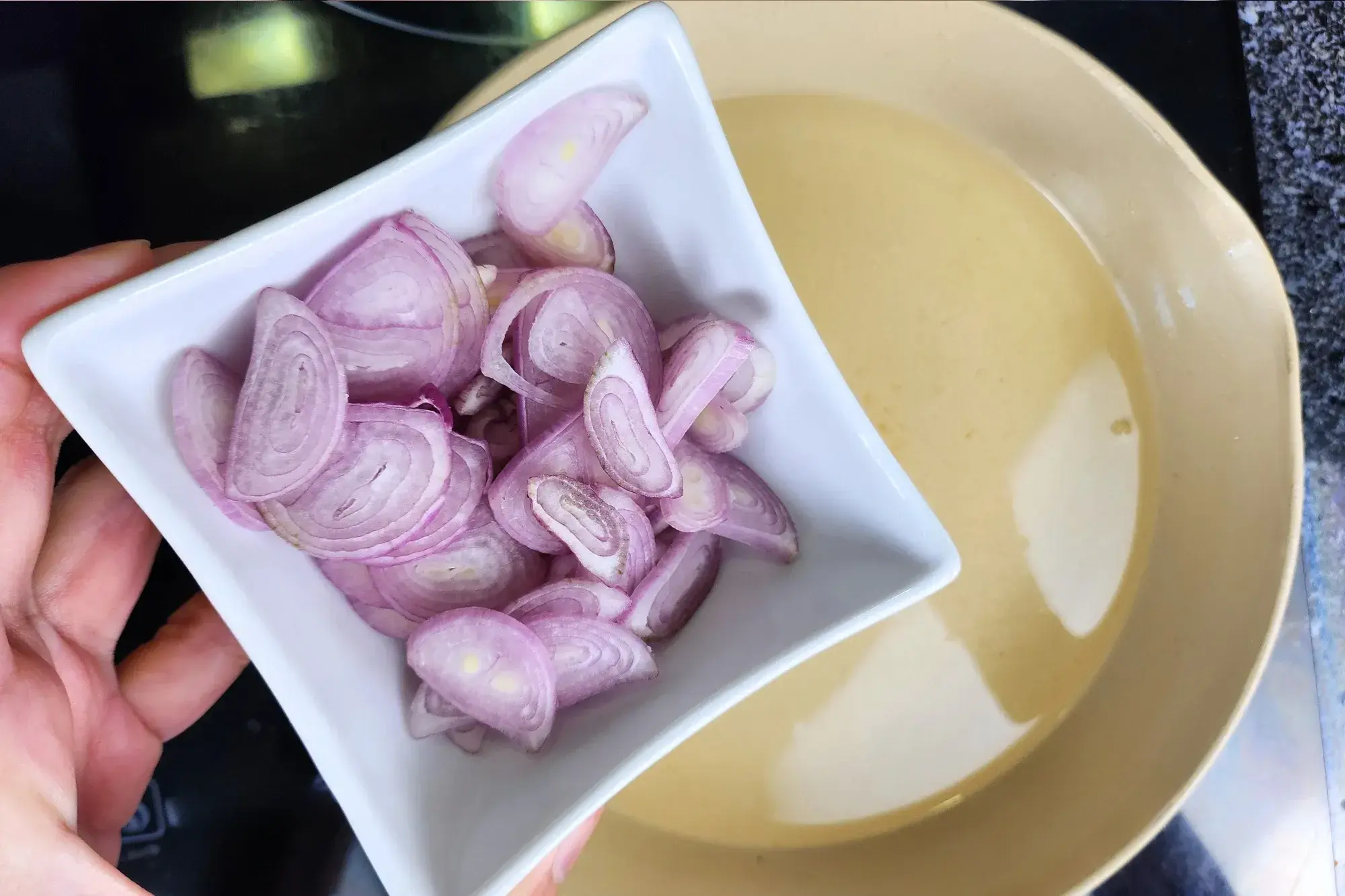 A hand is holding a bowl of sliced shallots above a sauce pan.