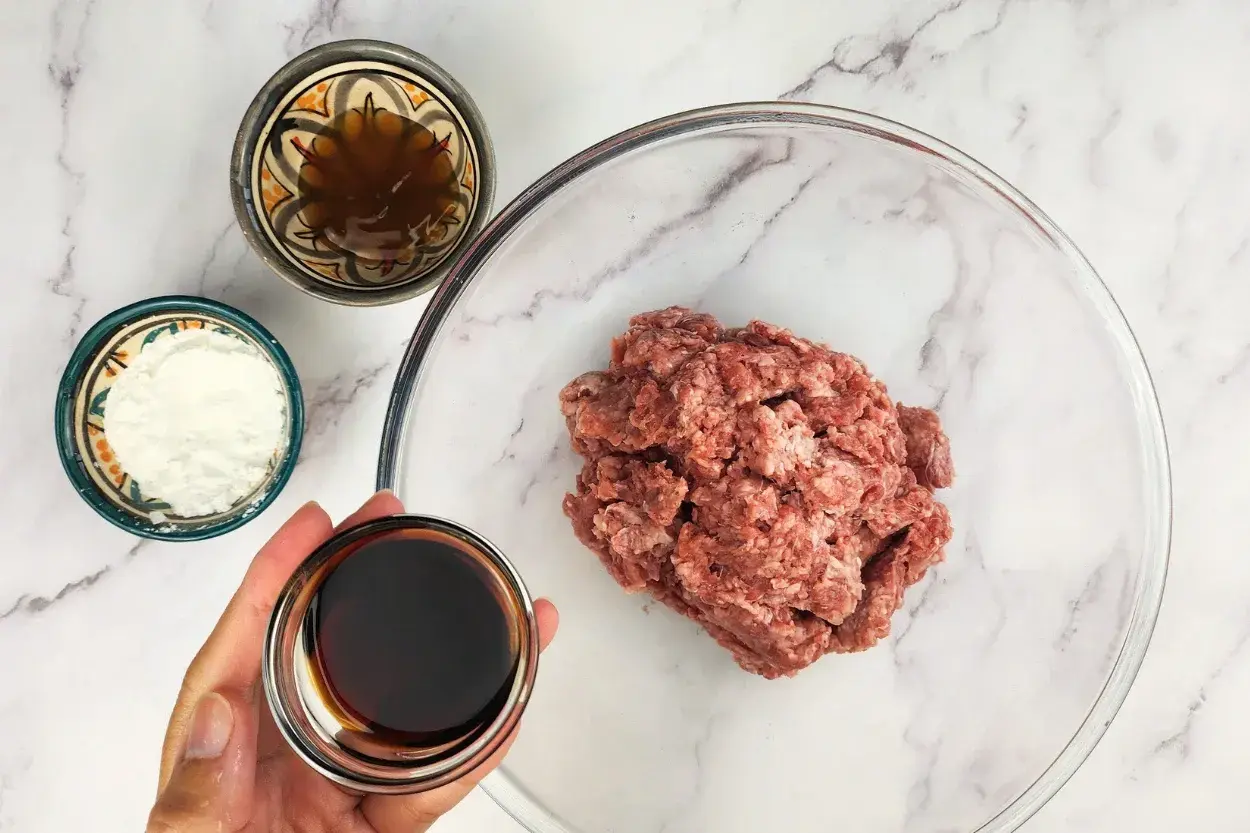 A hand is holding a bowl of oyster sauce above a mixing bowl with ground beef.