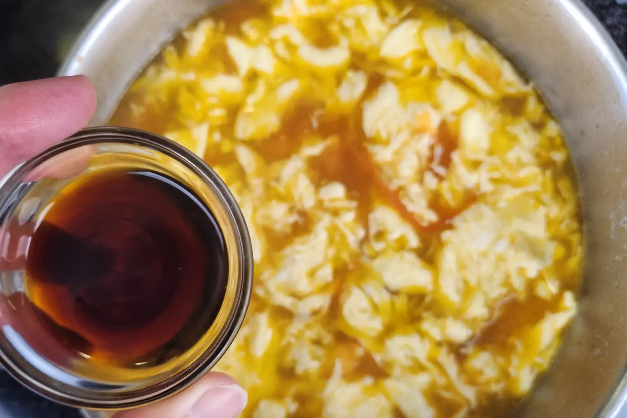 A hand is holding a bowl of soy sauce is above Tomato Egg Drop Soup.