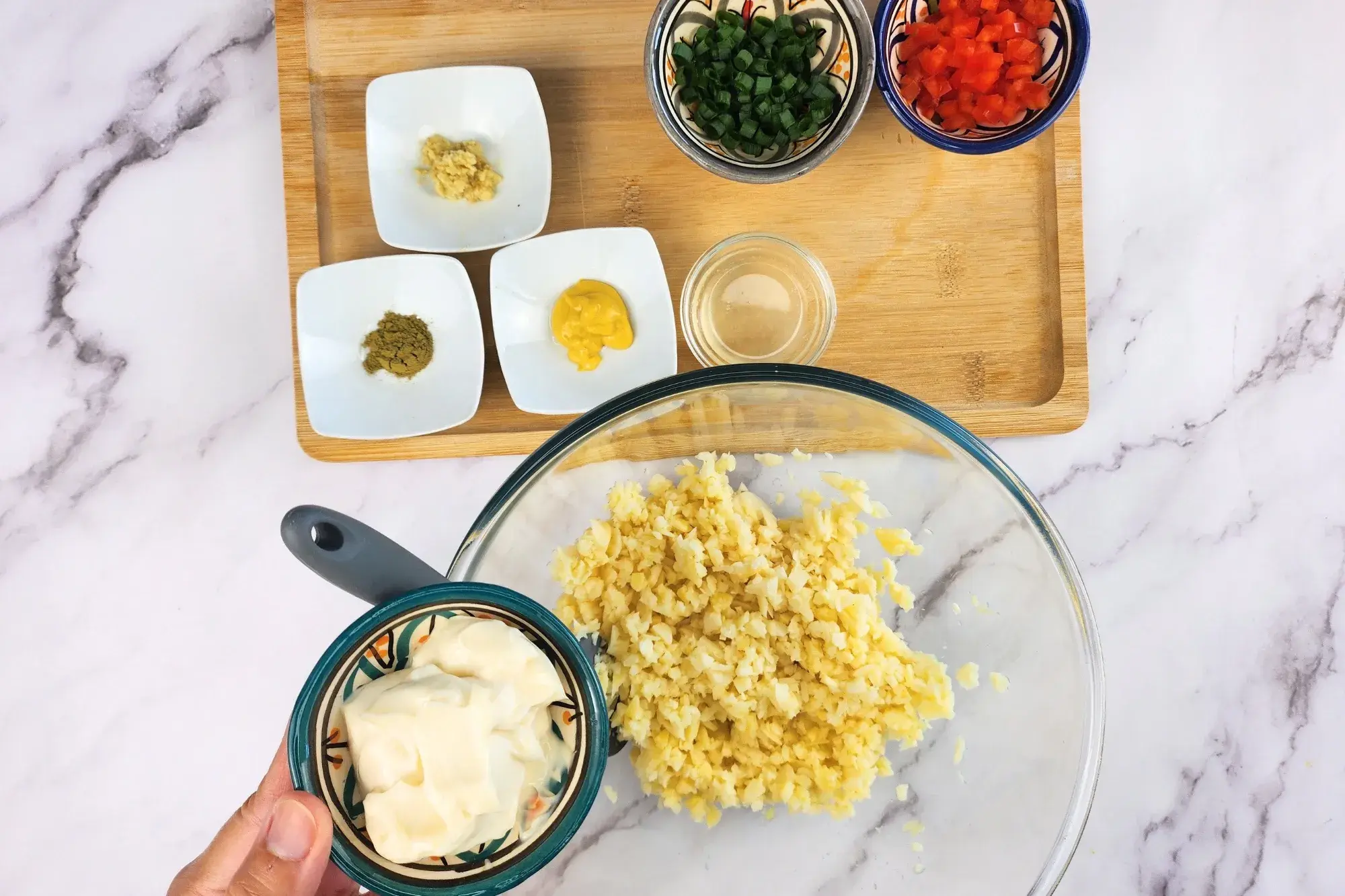 A hand is holding a bowl of vegan mayo above the mixing bowl.