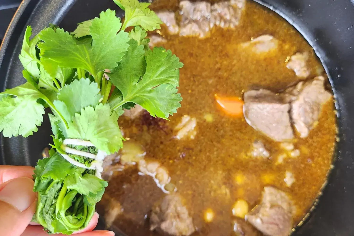 A hand is holding a bundle of cilantro above a pot with meat.
