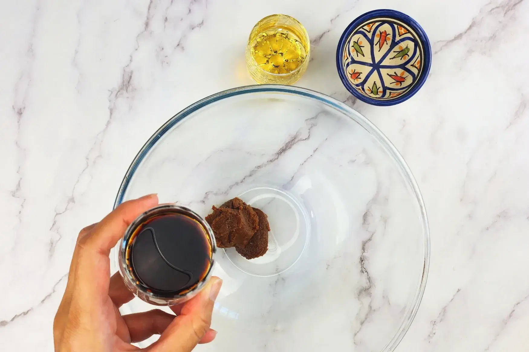 A hand is holding a glass bowl of soy sauce above a mixing bowl.