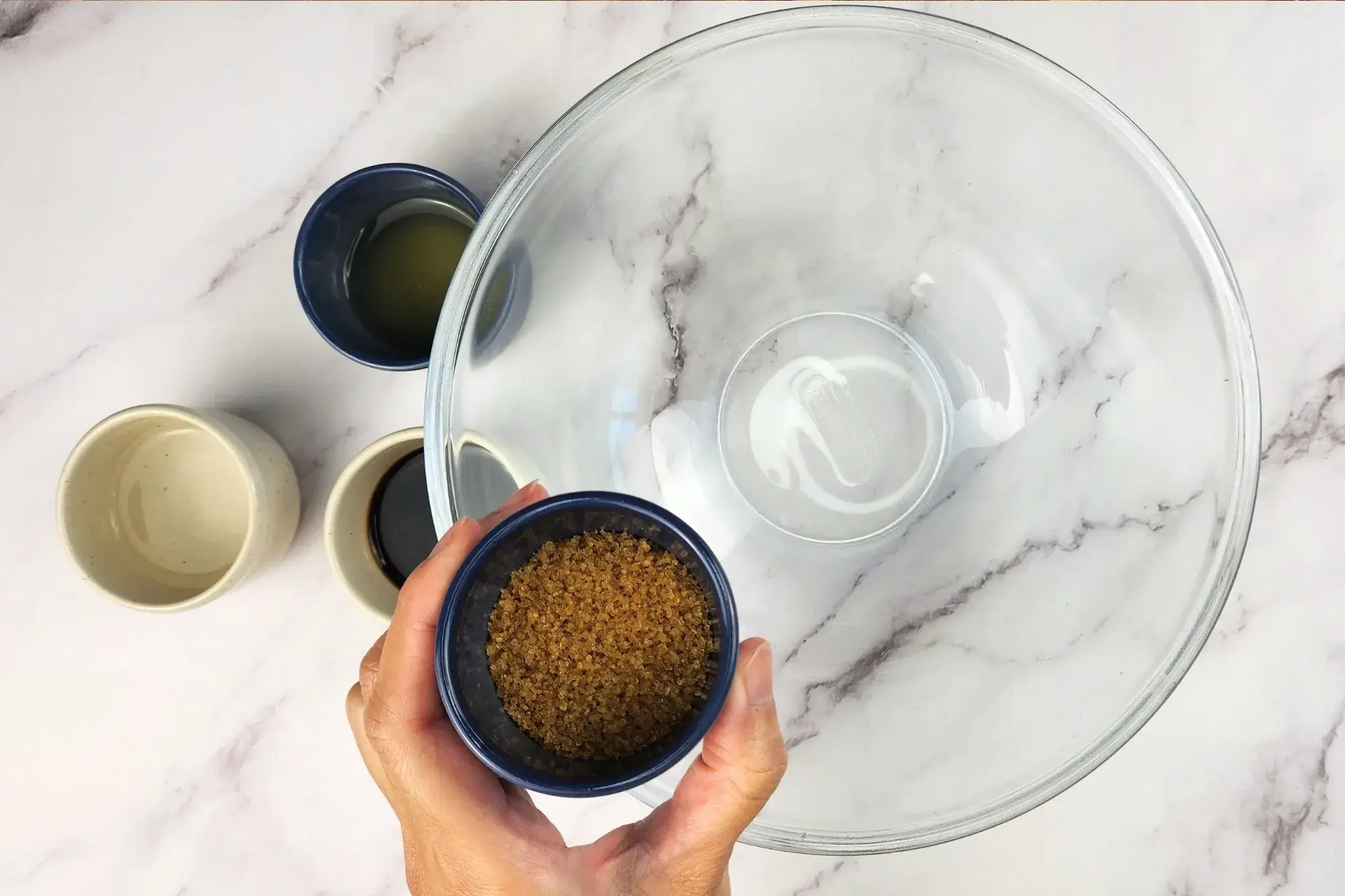 A hand is holding a small bowl of brown sugar above a mixing bowl.