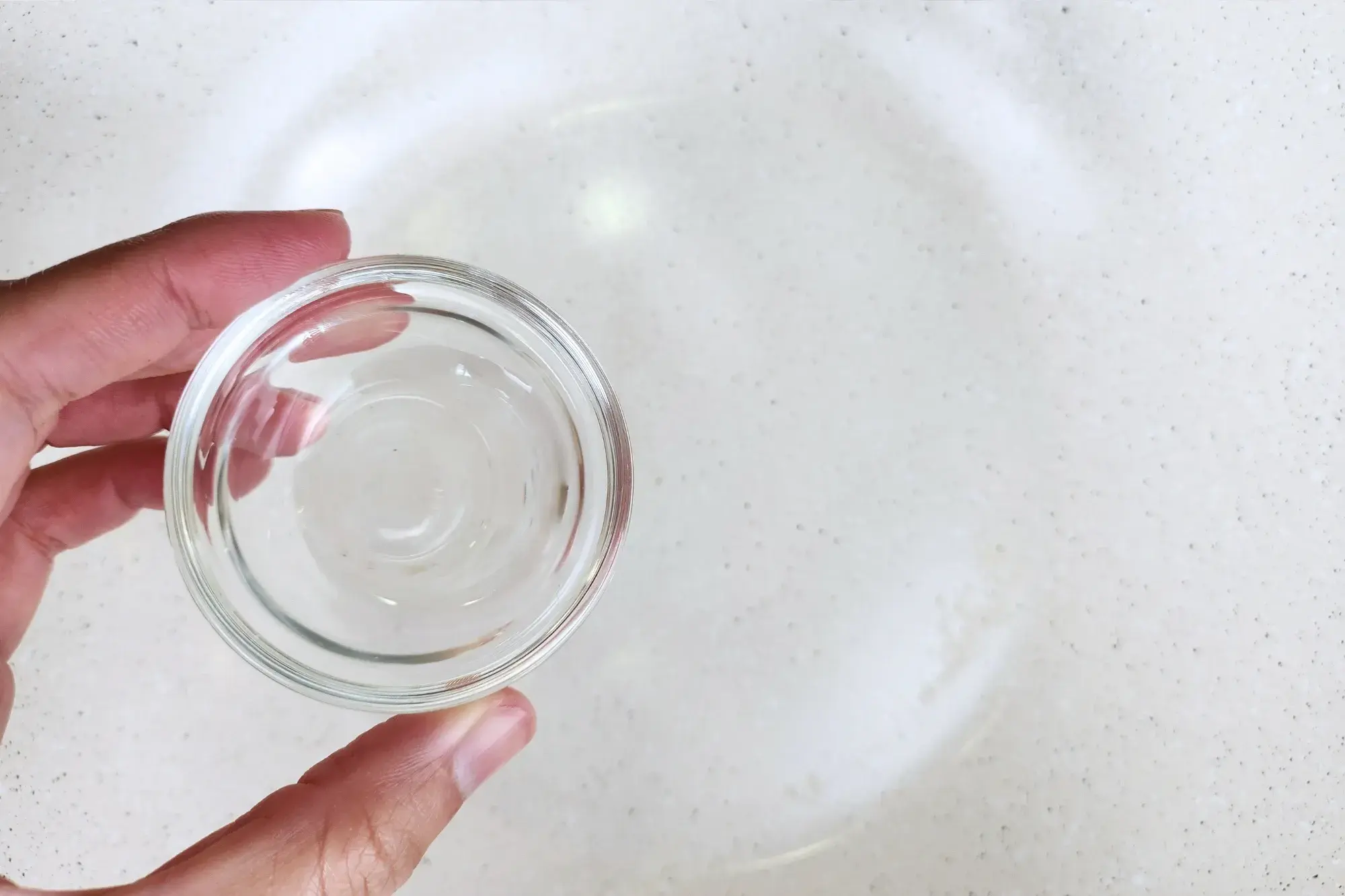 A hand is holding a small bowl of cooking oil above a wok