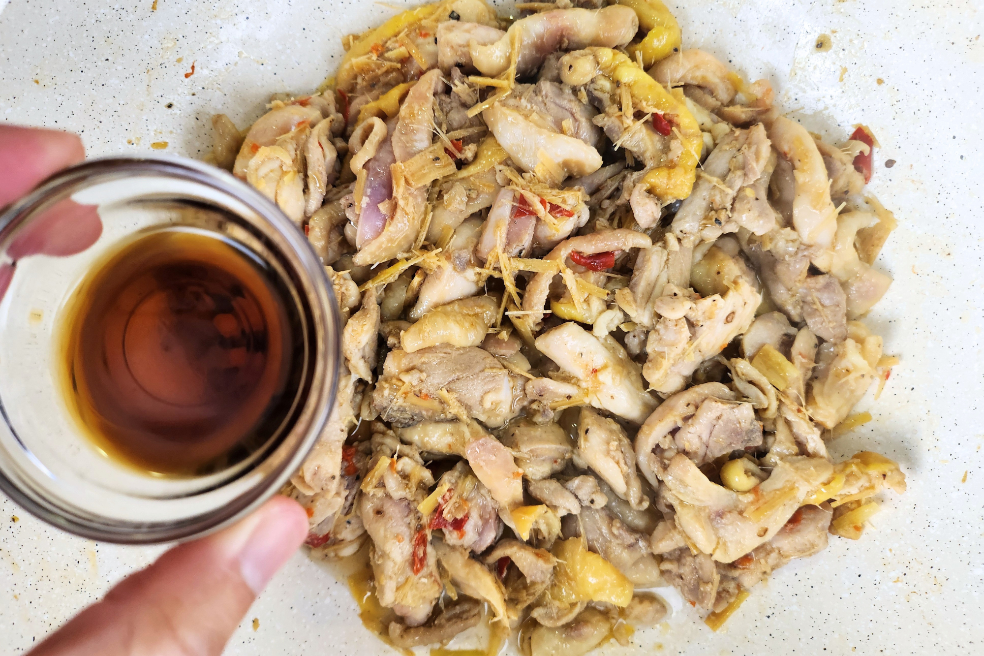 A hand is holding a small bowl of fish sauce is above a wok of cooked chicken.