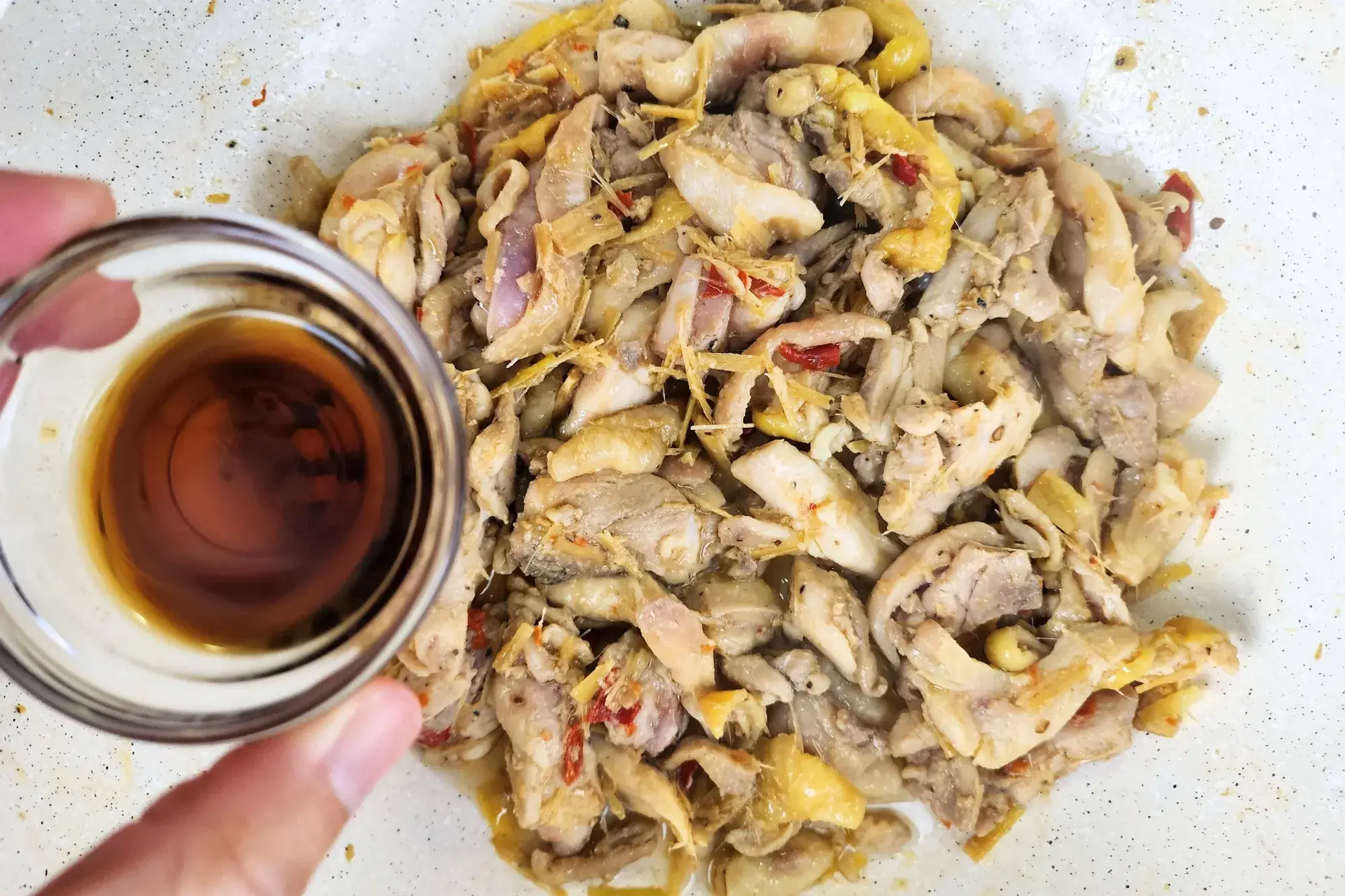 A hand is holding a small bowl of fish sauce is above a wok of cooked chicken.