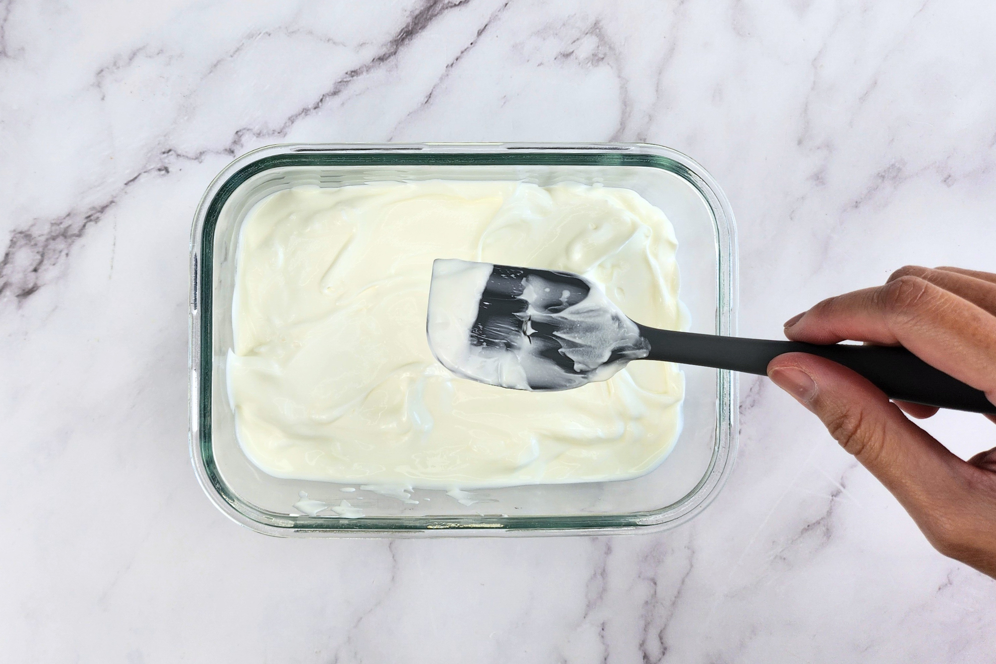 A hand is holding a spatula above a bowl of yogurt.