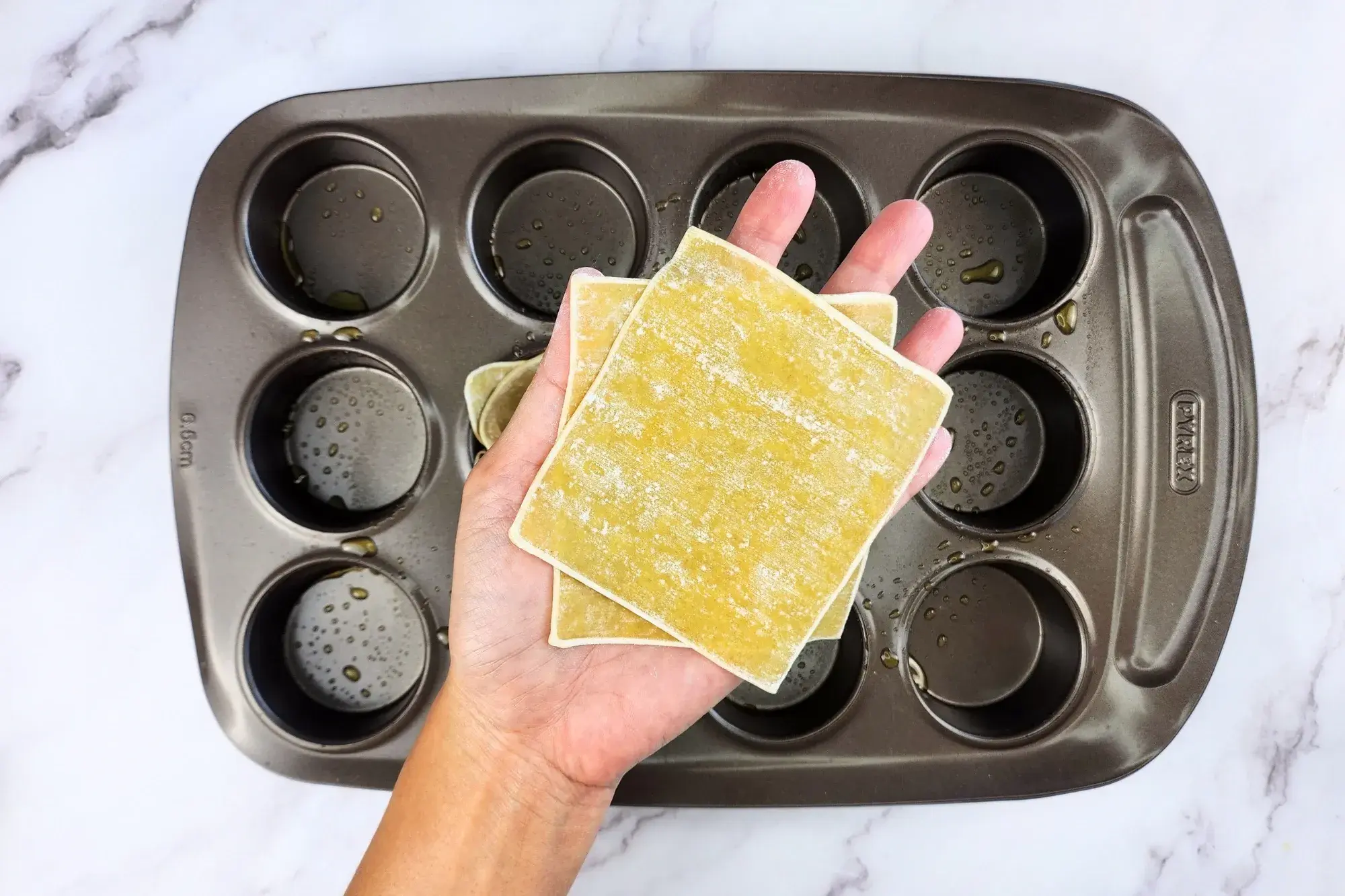 A hand is holding two wonton wrappers above the muffin tray.