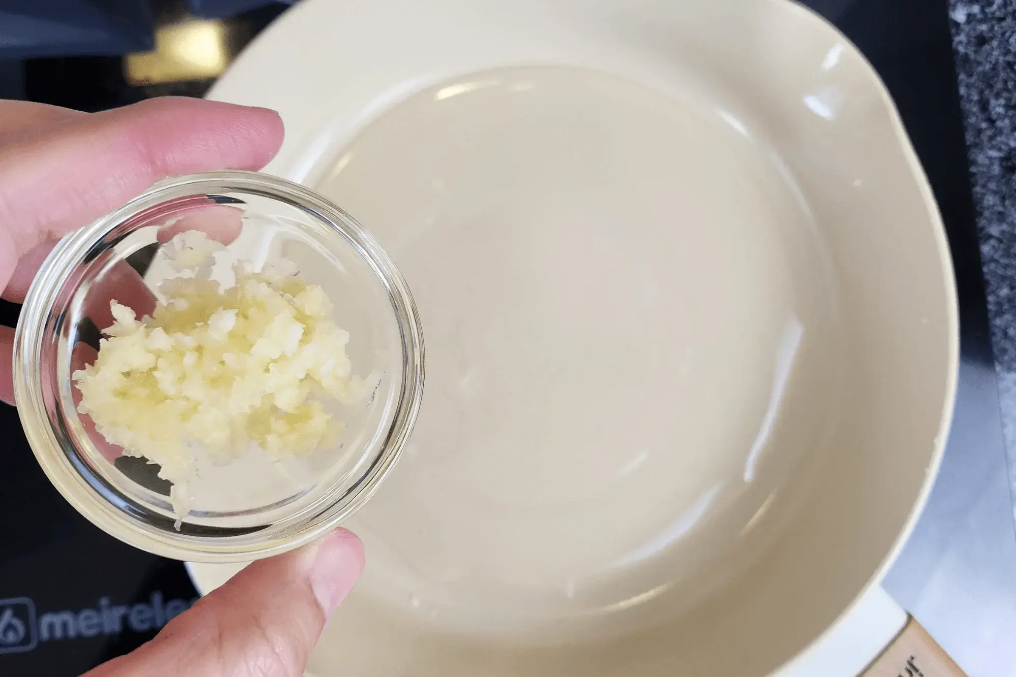 a hand holding a bowl of minced garlic with the sauce pan