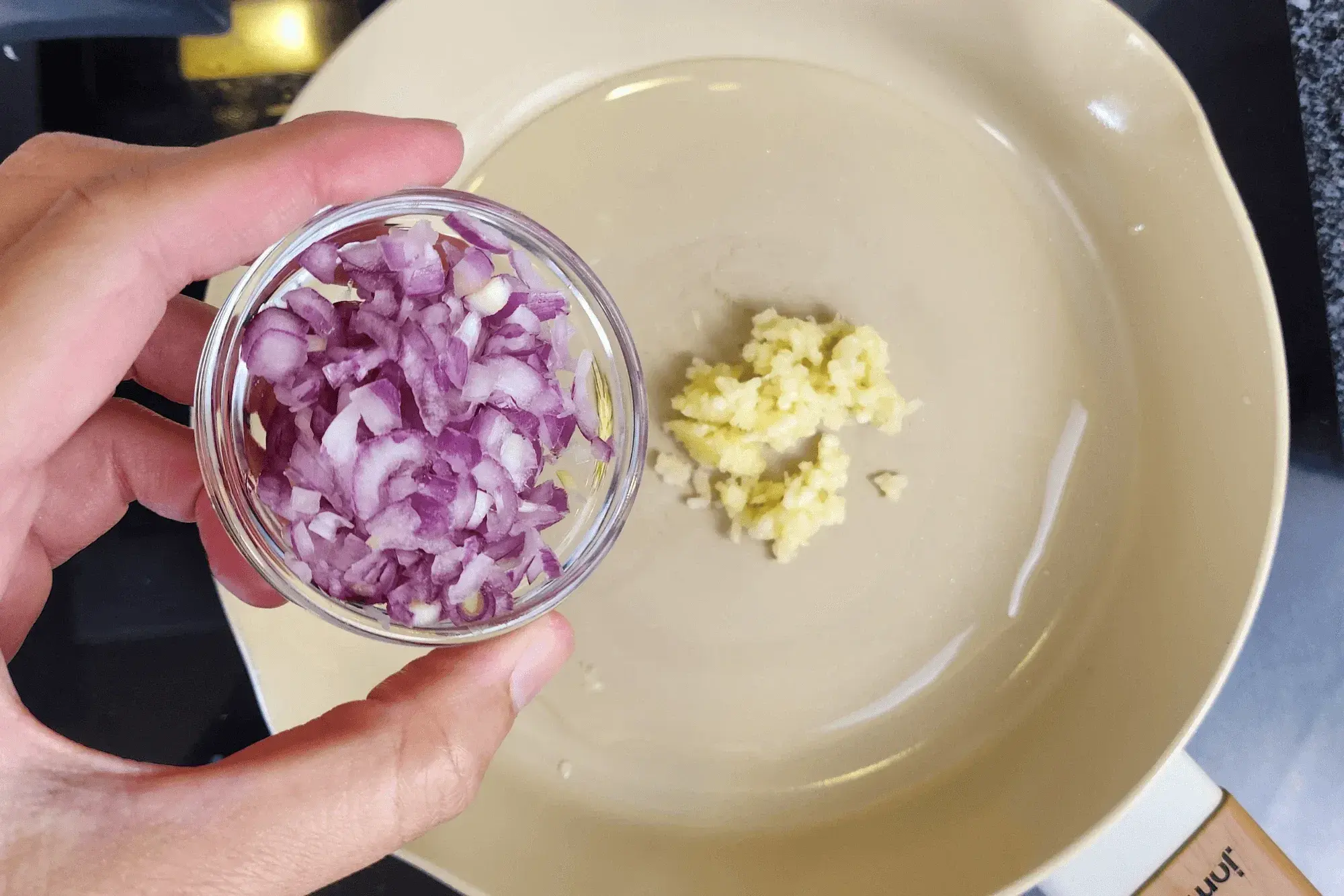 a hand holding a bowl of minced shallots with the sauce pan