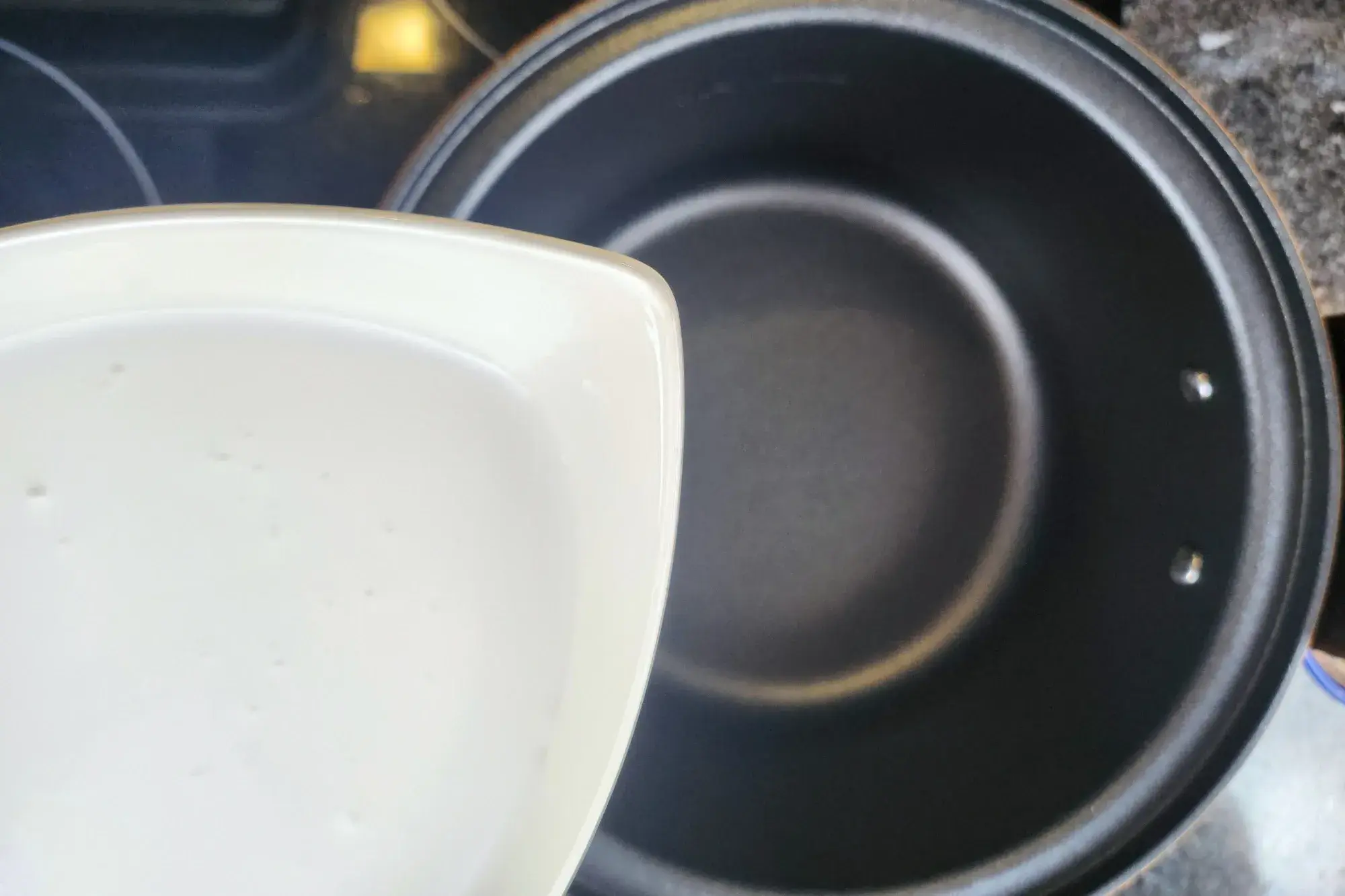 Coconut milk is being poured from a bowl into an empty black pot on a stovetop.