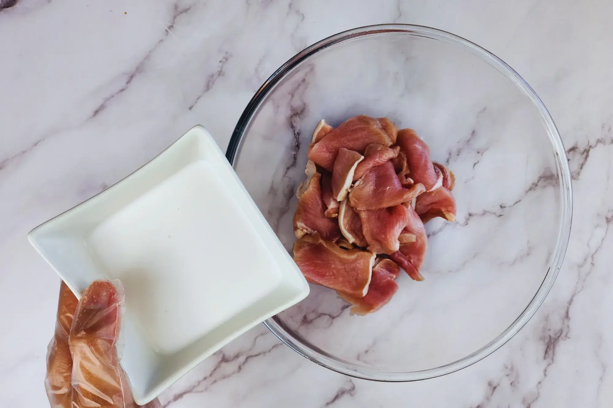 Pork slices in a mixing bowl and a hand holding of bowl of coconut milk
