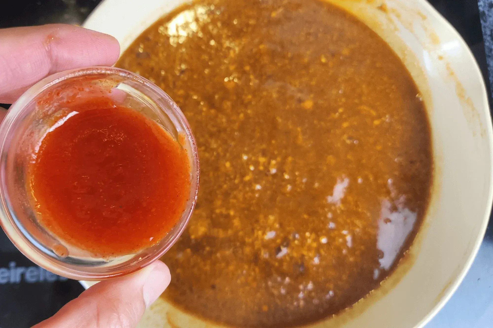 a hand holding a bowl of Sriracha sauce with the sauce pan