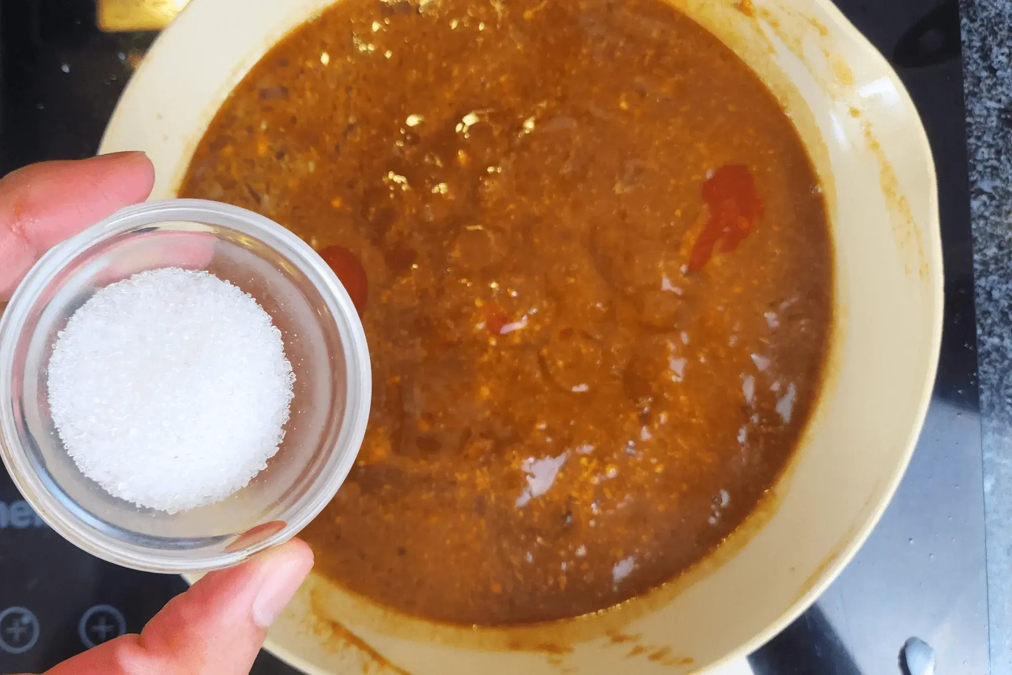 a hand holding a bowl of suagr with the sauce pan