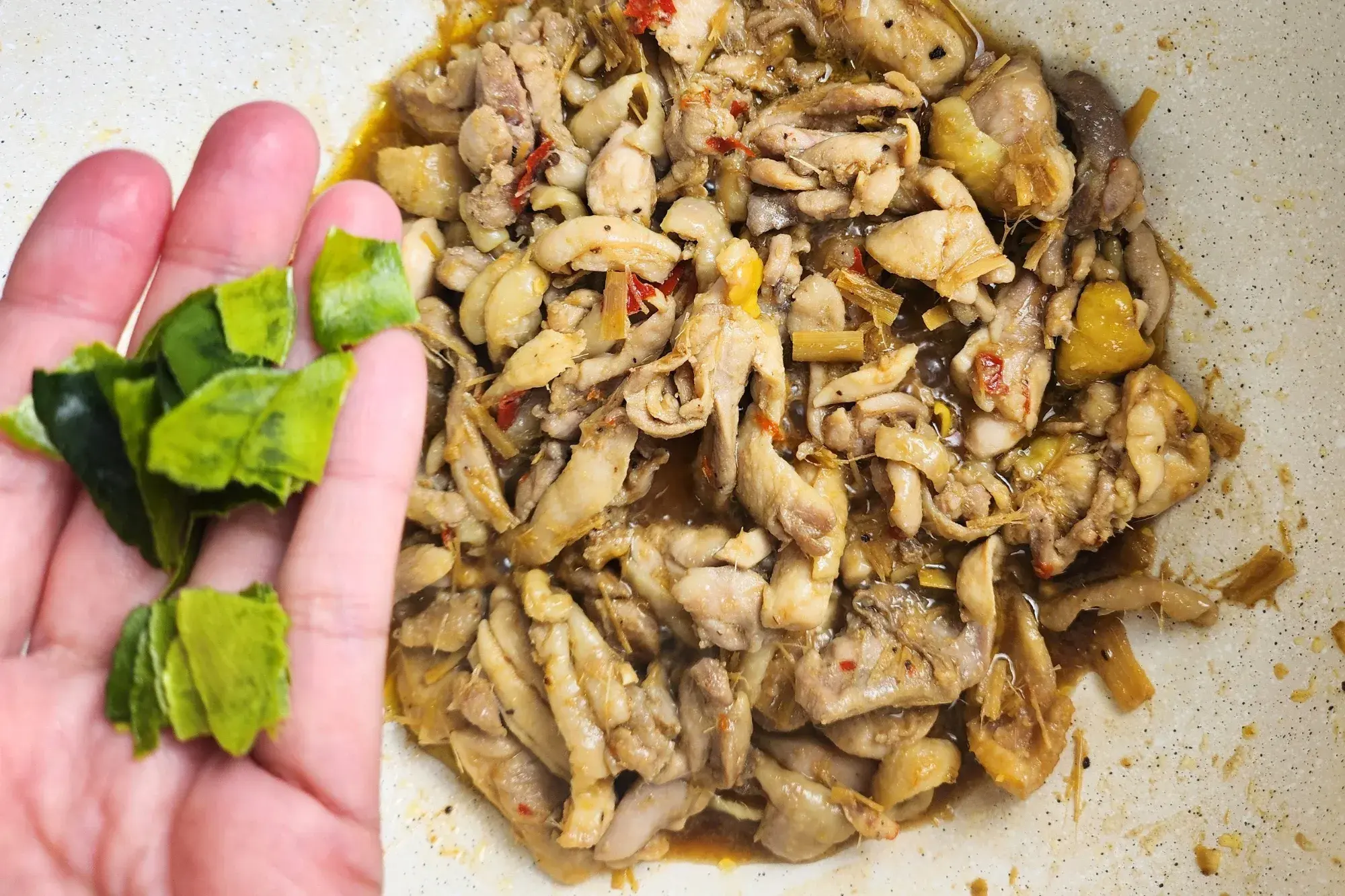 Tear kaffir lime leaves are in a hand above a wok of cooked chicken.