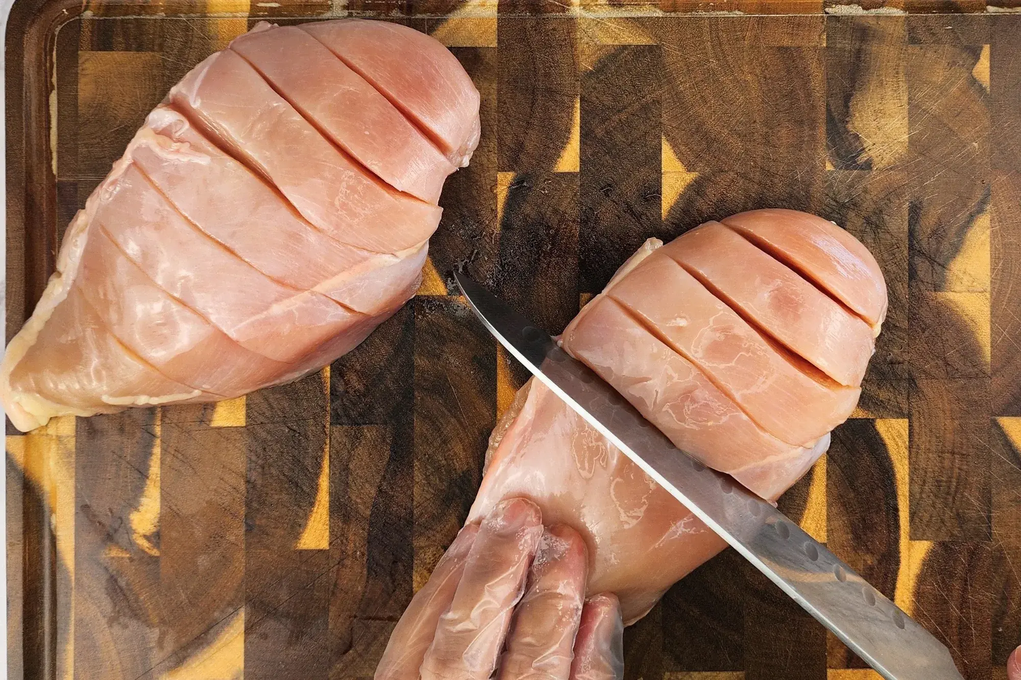 Two cut chicken breasts, a hand holding a knife cut a chicken breast