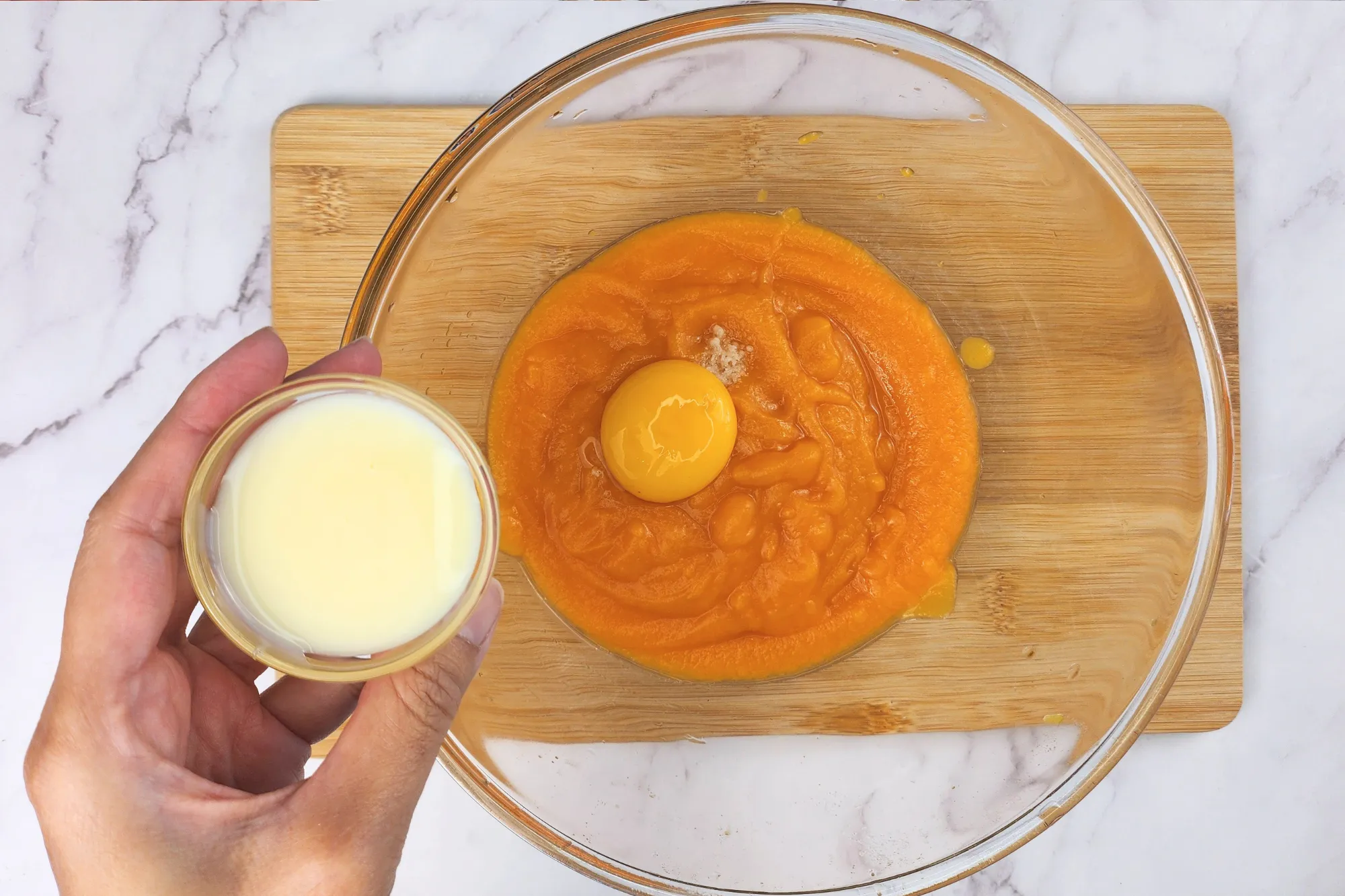 A hand is holding a bowl of condensed milk above a mixing bowl.