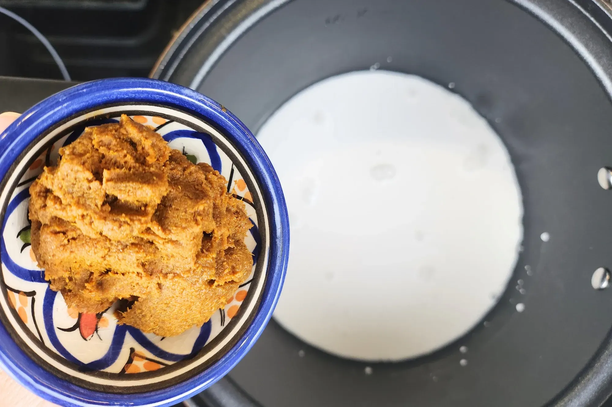 A bowl of Thai Yellow Curry Paste is held above a pot containing coconut milk on a stovetop.