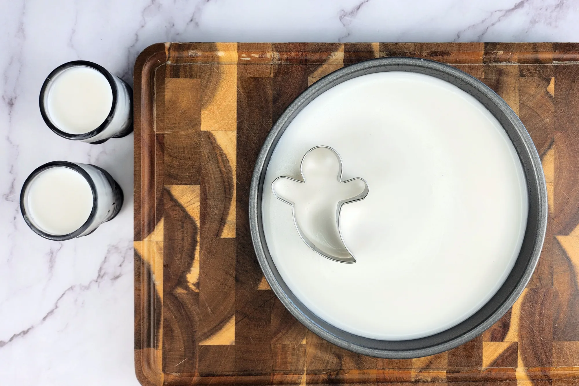 A round tray with white liquid on chopping board and a cookie cutter and two glasses shot