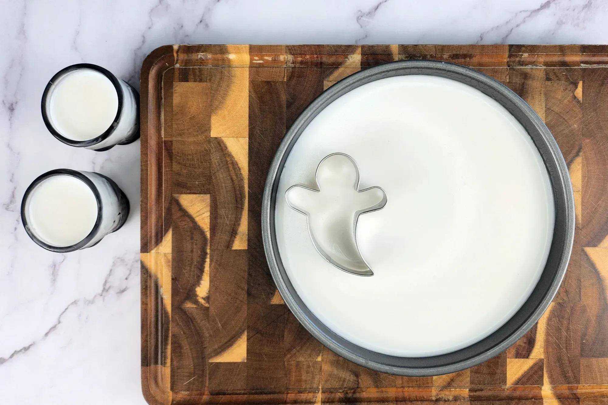 A round tray with white liquid on chopping board and a cookie cutter and two glasses shot