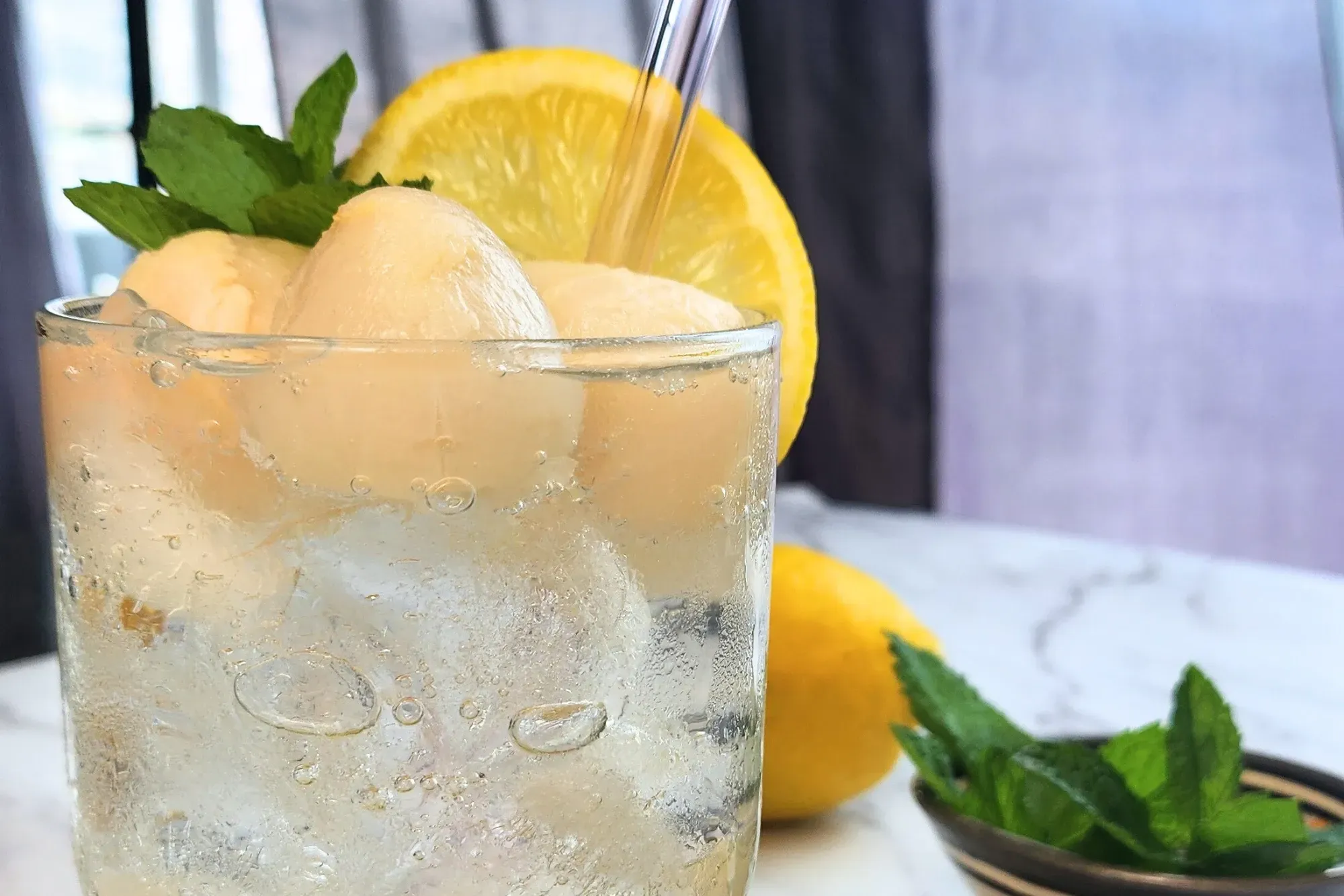 A glass of lychee lemonade is topping with lychees, mint and a slice of lemon with a straw.