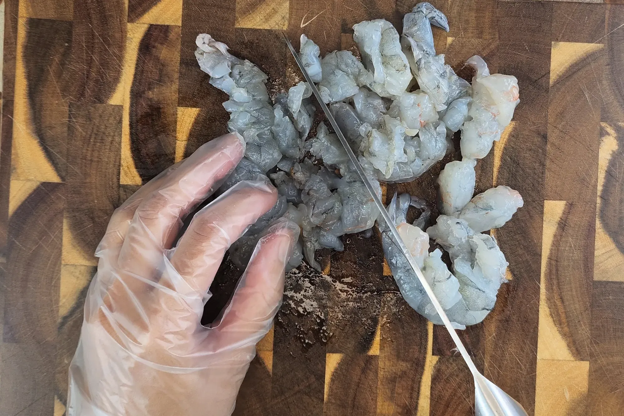 A hand above chopping board with a knife is copping raw shrimp